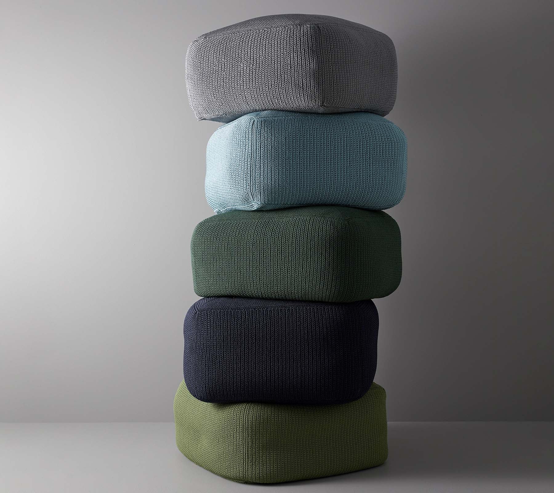 A stacked arrangement of five cushions in various colors, including grey, light blue, green, dark blue, and olive green.