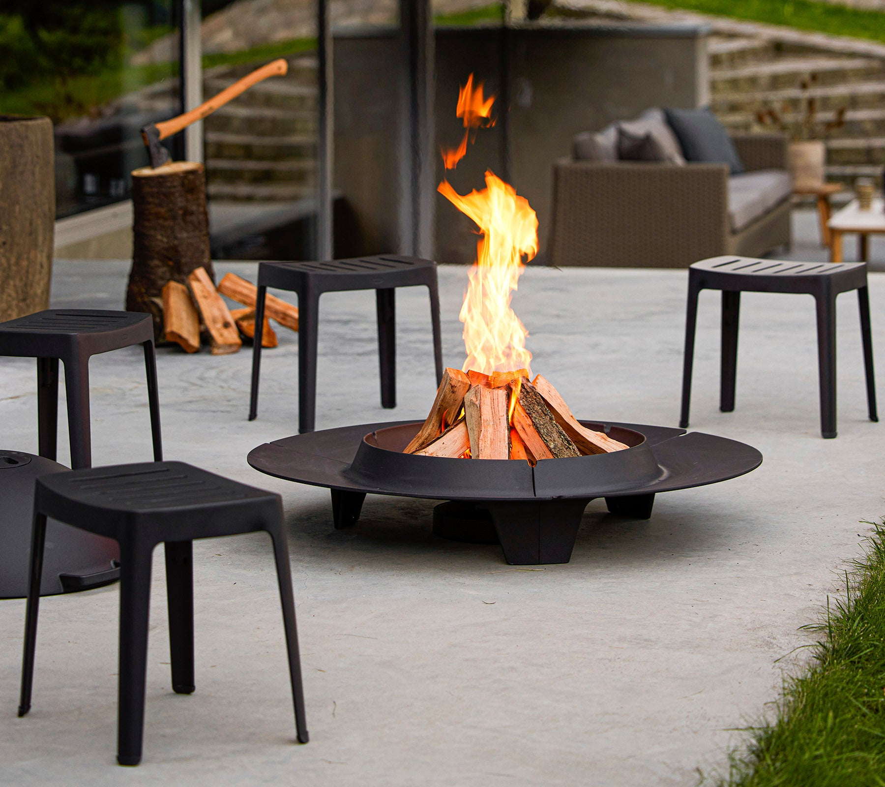 A fire pit surrounded by minimalist black chairs, with a vibrant flame and stacks of firewood nearby.