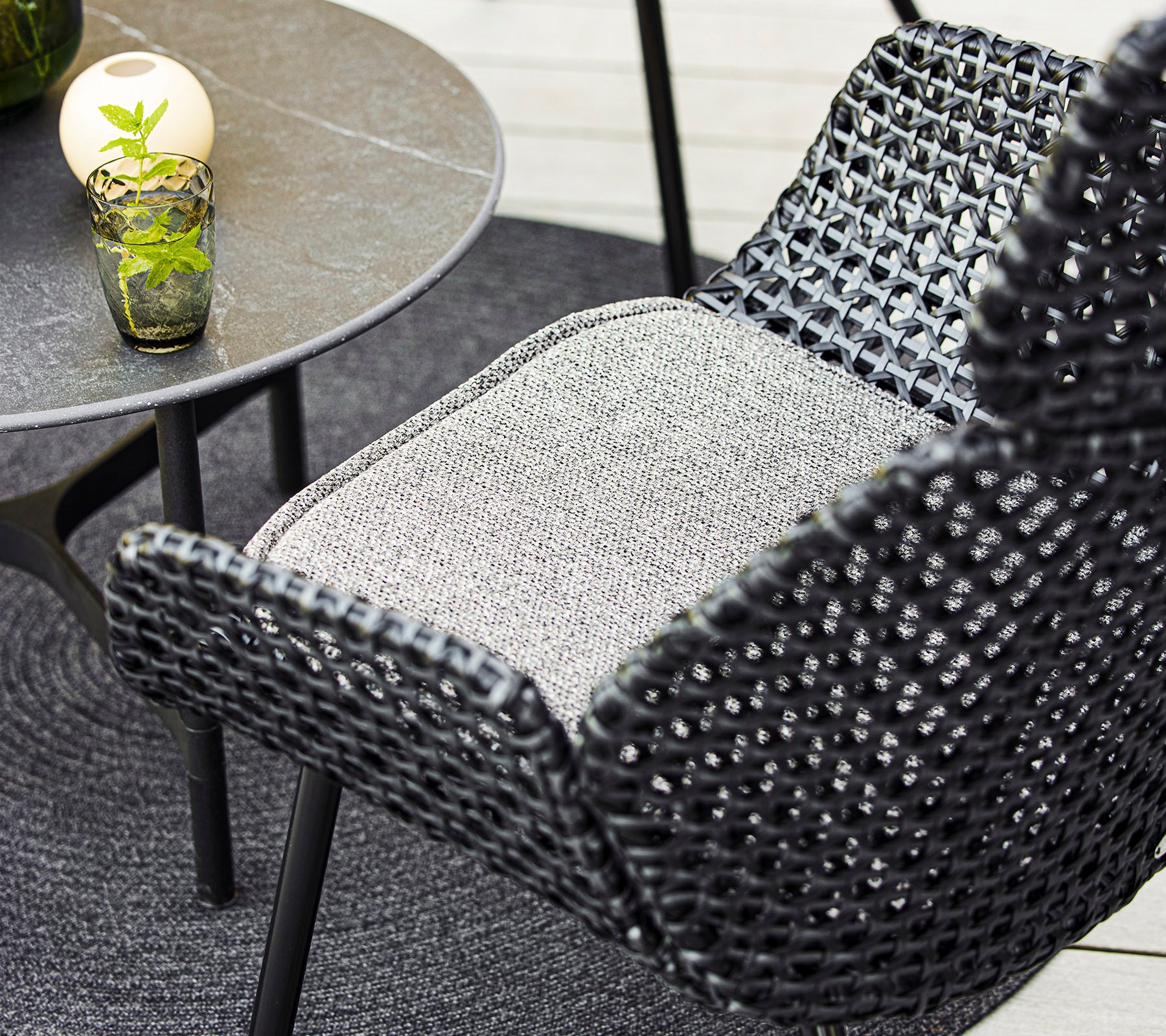 A close-up view of a stylish woven chair and a small table with a decorative glass and a plant, paired with a cozy circular mat.