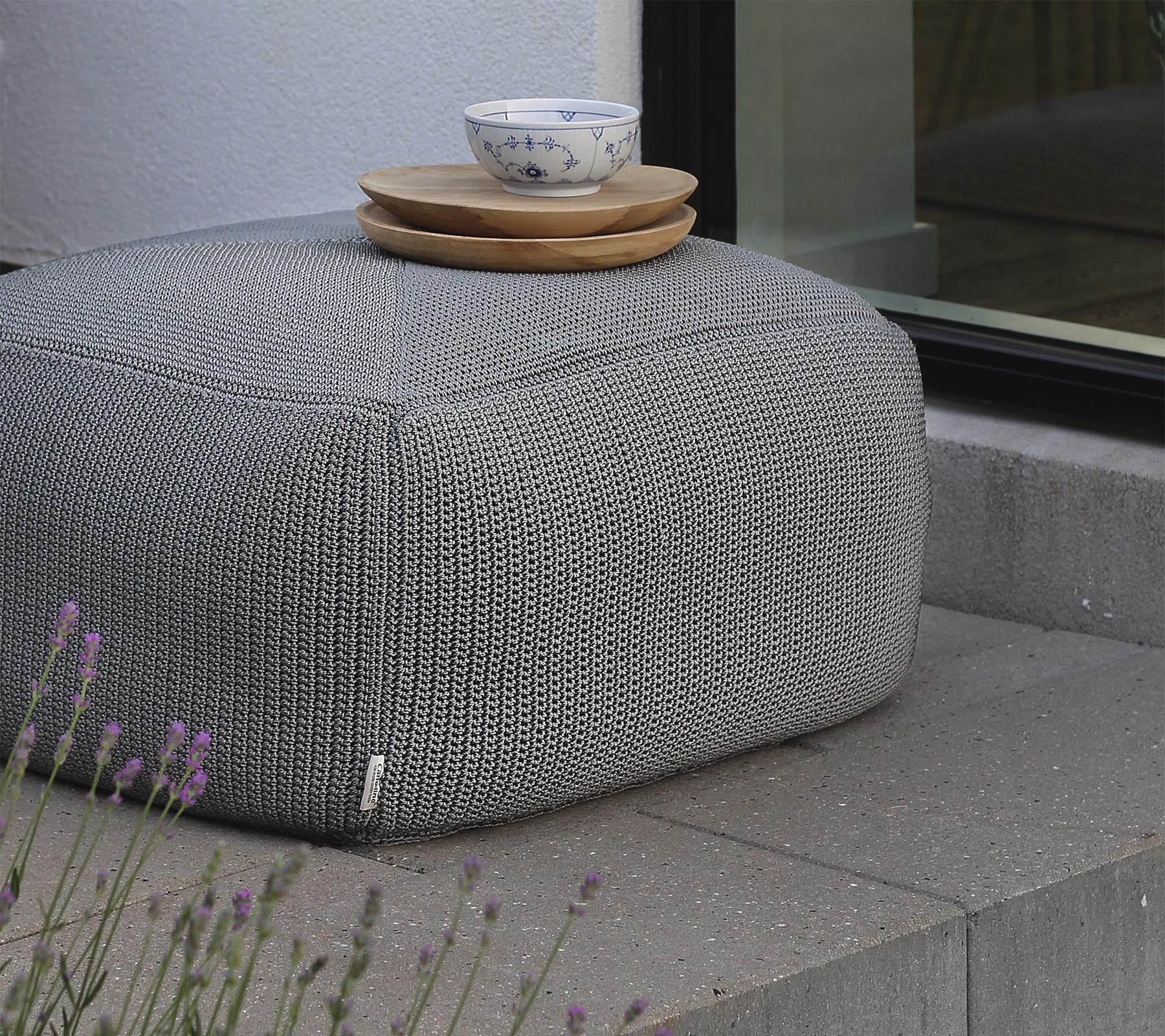 A textured gray pouf topped with wooden plates and a ceramic bowl, positioned near a window and surrounded by lavender.