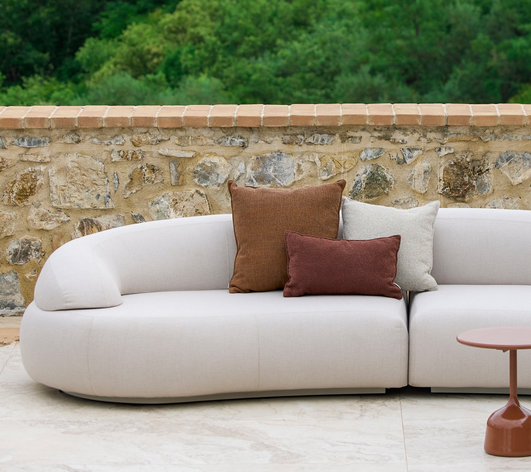 A curved sofa adorned with Rise cushions in earthy tones alongside a stylish Glaze side table, set against a stone backdrop.