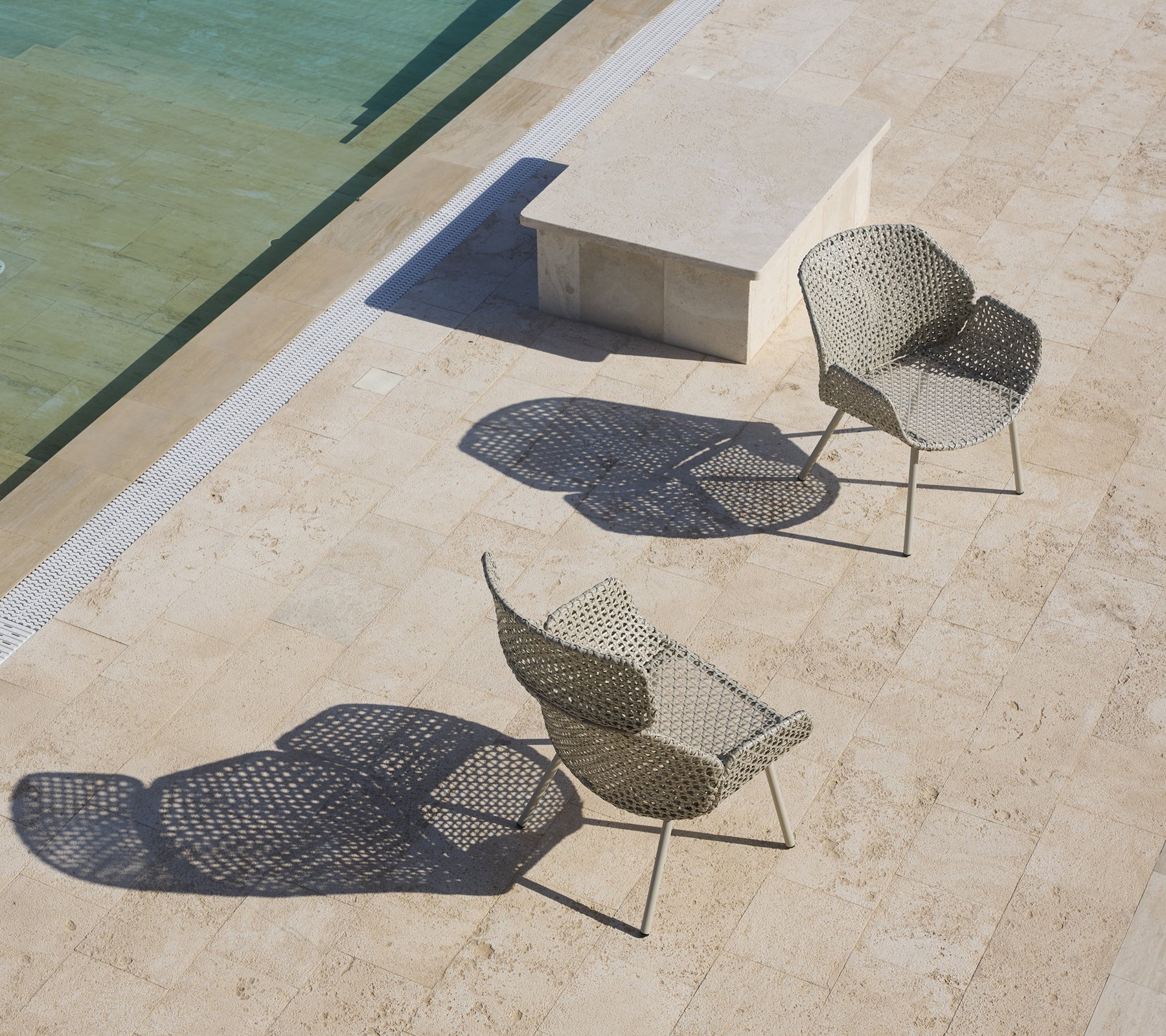 Two stylish lounge chairs with unique designs casting shadows, positioned near a calm water surface and a stone table.