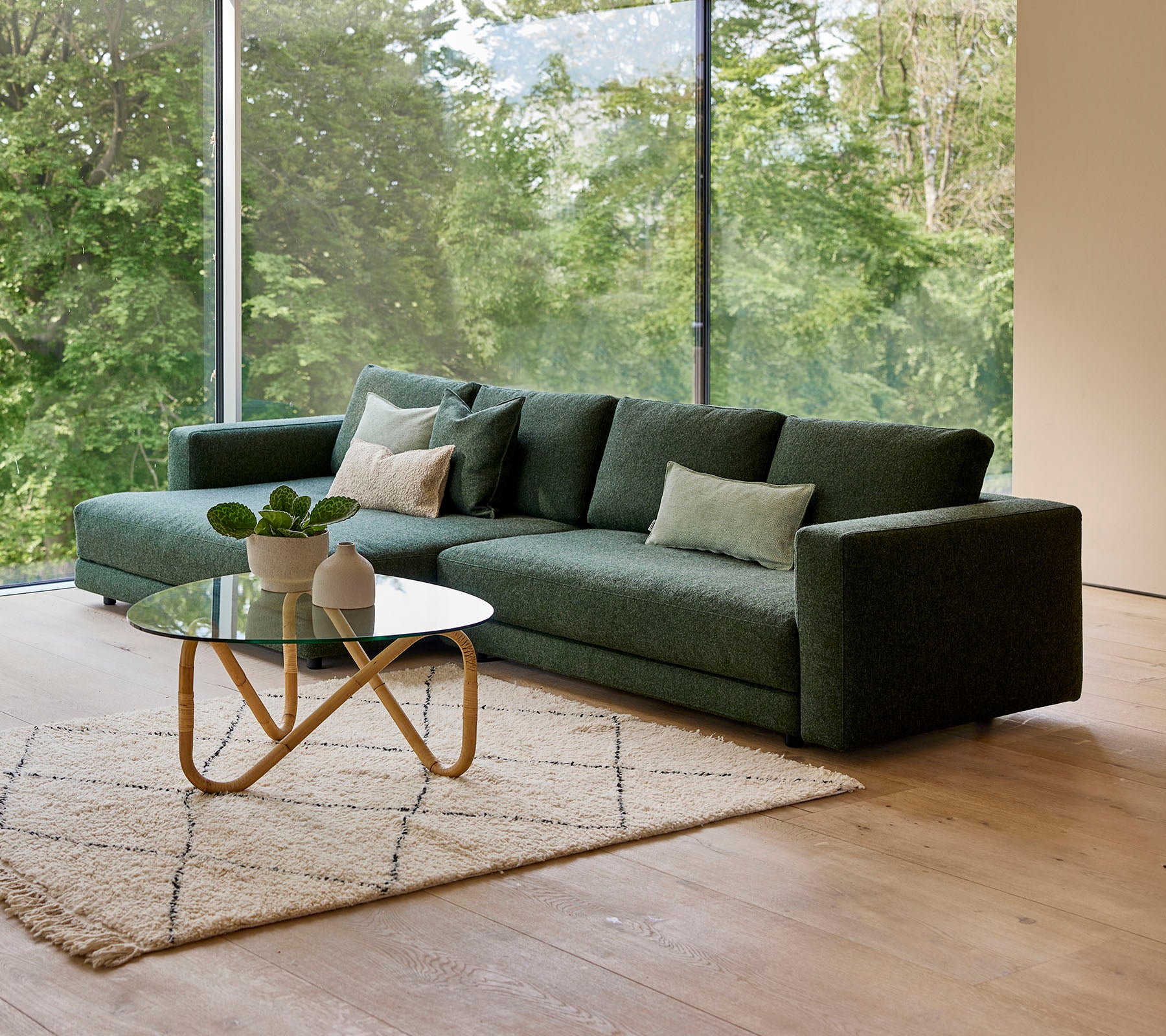 A modern green sofa with pillows, accompanied by a round coffee table and a decorative rug, set against a backdrop of greenery.