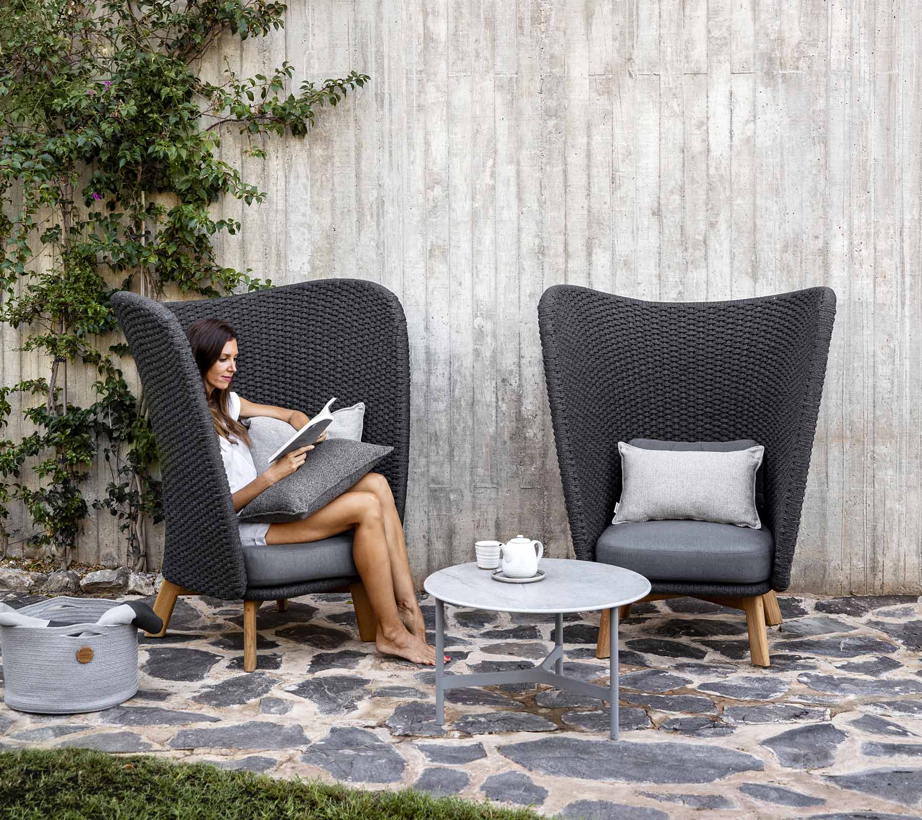 A woman reads comfortably while seated in a stylish wicker chair beside a small table, surrounded by greenery and smooth stones.