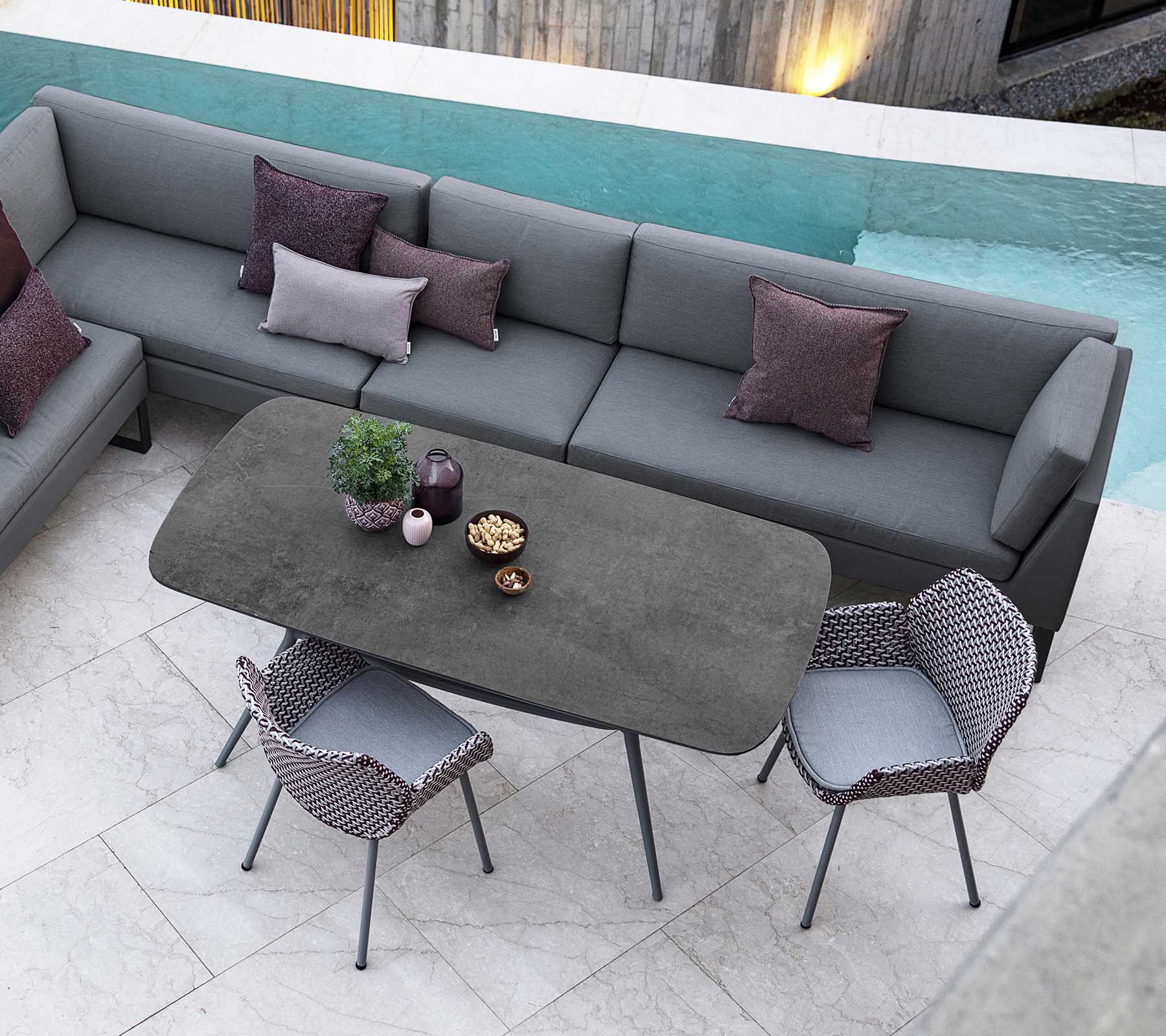 Modern outdoor seating area featuring a sectional sofa with pillows, a sleek rectangular table, and stylish chairs, complemented by a pool view.