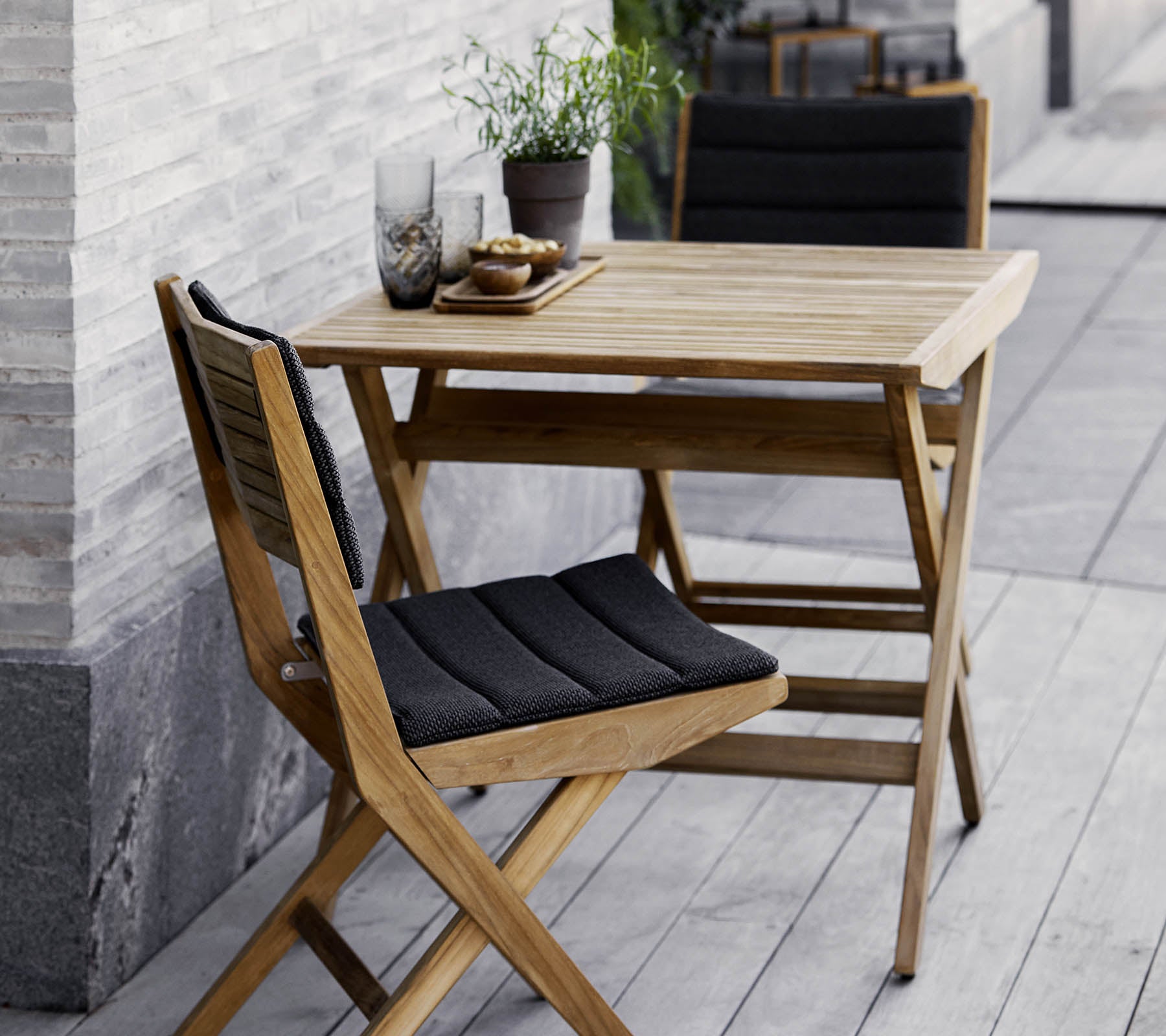 Wooden table and chair set featuring black cushions, adorned with potted plants and decorative glassware. Perfect for a cozy setting.