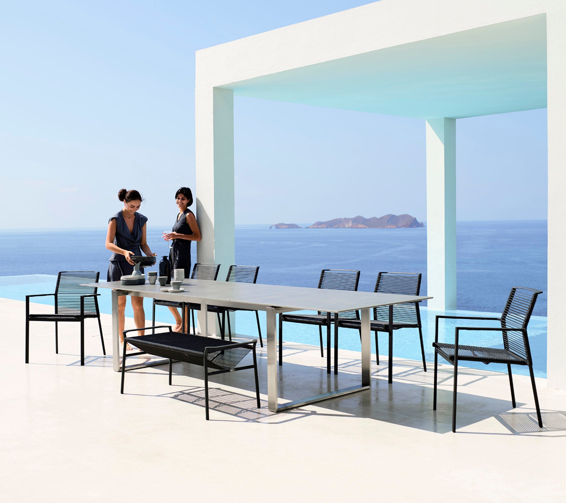 Two women prepare a dining table set with sleek black chairs against a backdrop of a serene blue sea and sky.