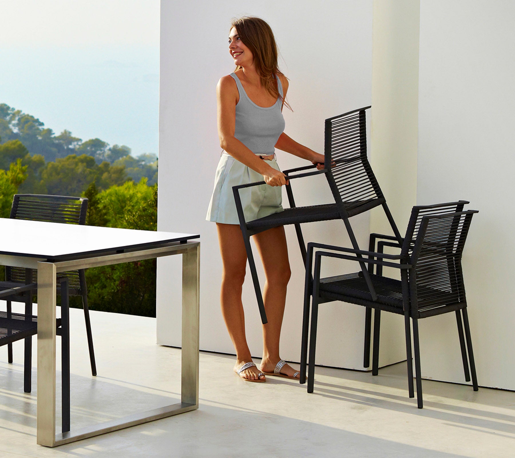A woman in a light tank top and shorts carries stacked black chairs next to a sleek table, set against a blurred natural backdrop.