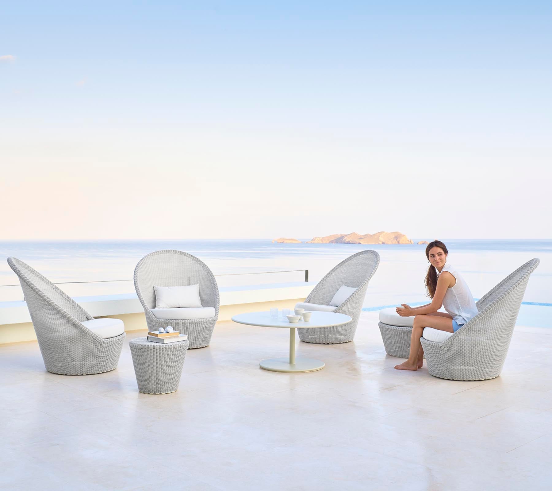 A stylish seating arrangement featuring elegant chairs and a table, with a woman sitting gracefully by a serene water backdrop.