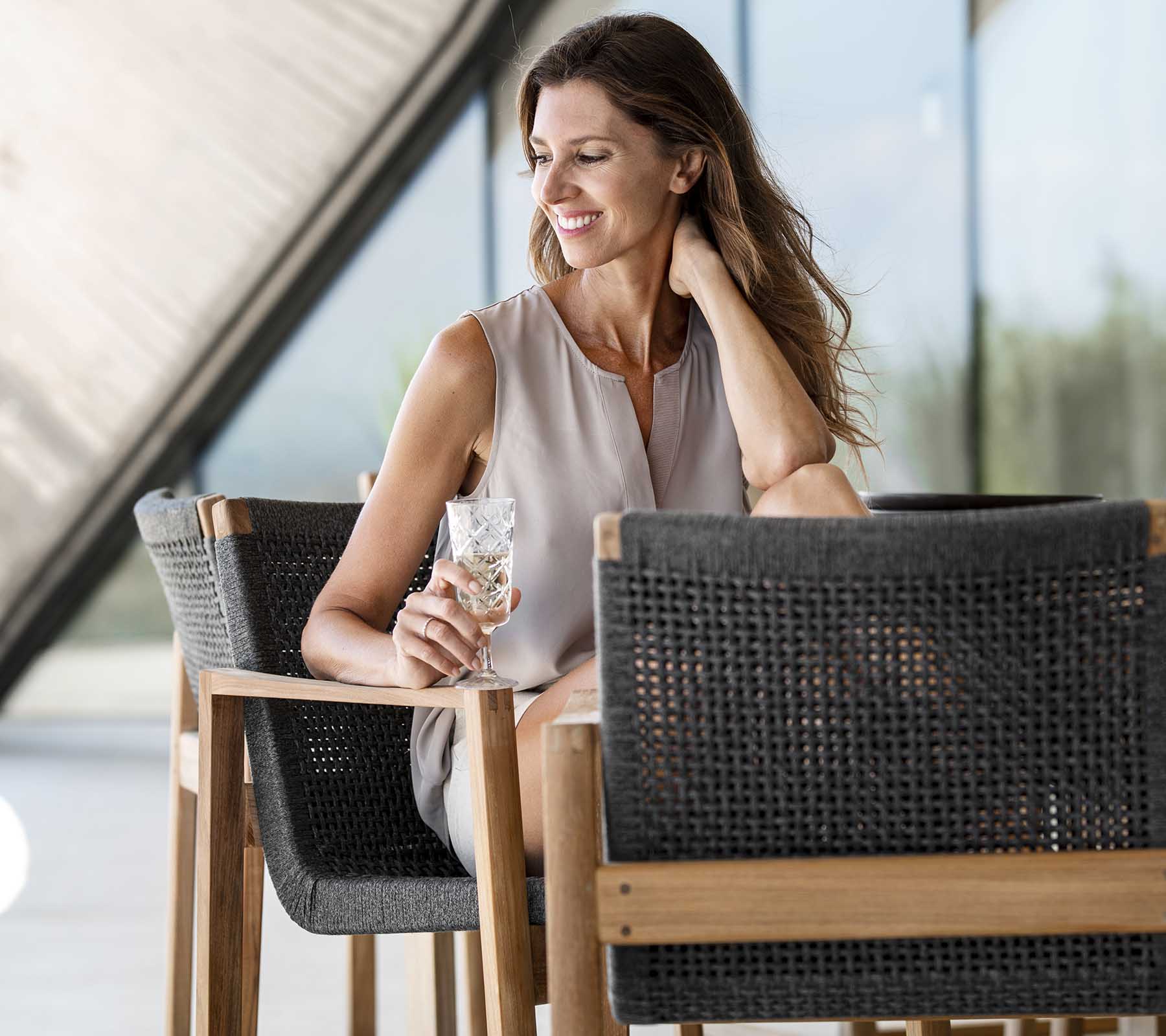 Smiling woman holding a drink while seated at a stylish chair, exuding a relaxed and elegant vibe. Bright and airy atmosphere.