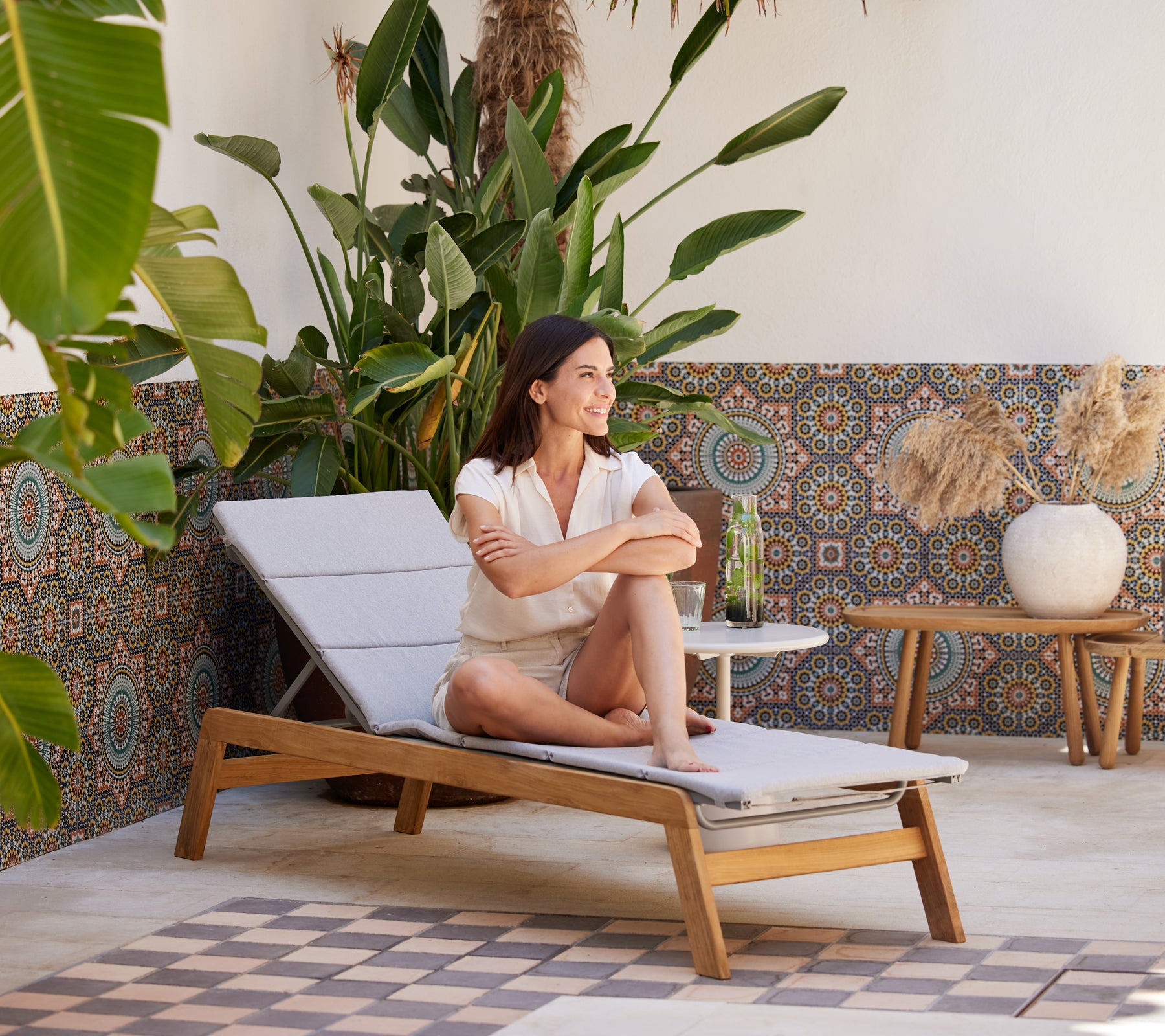 A woman relaxes on a chaise lounge surrounded by vibrant plants and patterned decor, enjoying a refreshing drink.