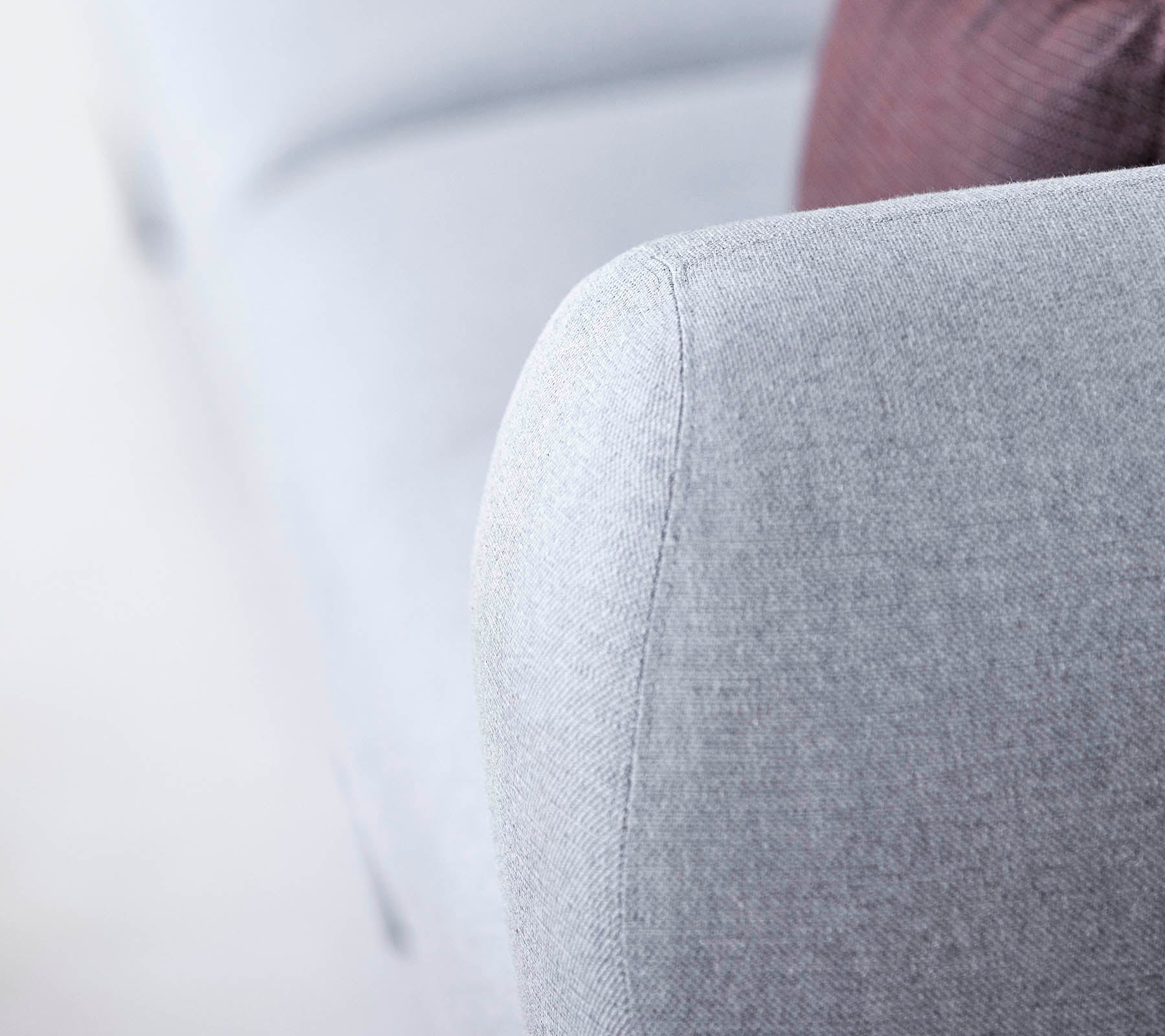 Close-up of a light gray sofa arm, featuring a textured fabric and a soft, rounded design, with a hint of a darker pillow in the background.