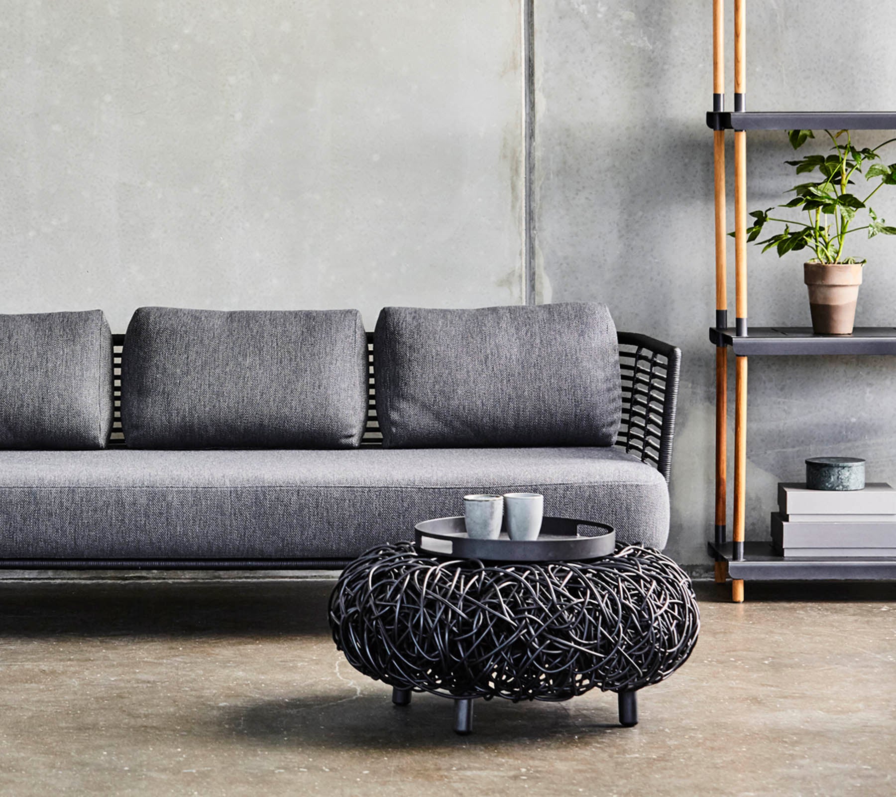 A modern gray sofa with cushions, paired with a circular black woven coffee table, and a minimalist shelf displaying plants and decor.