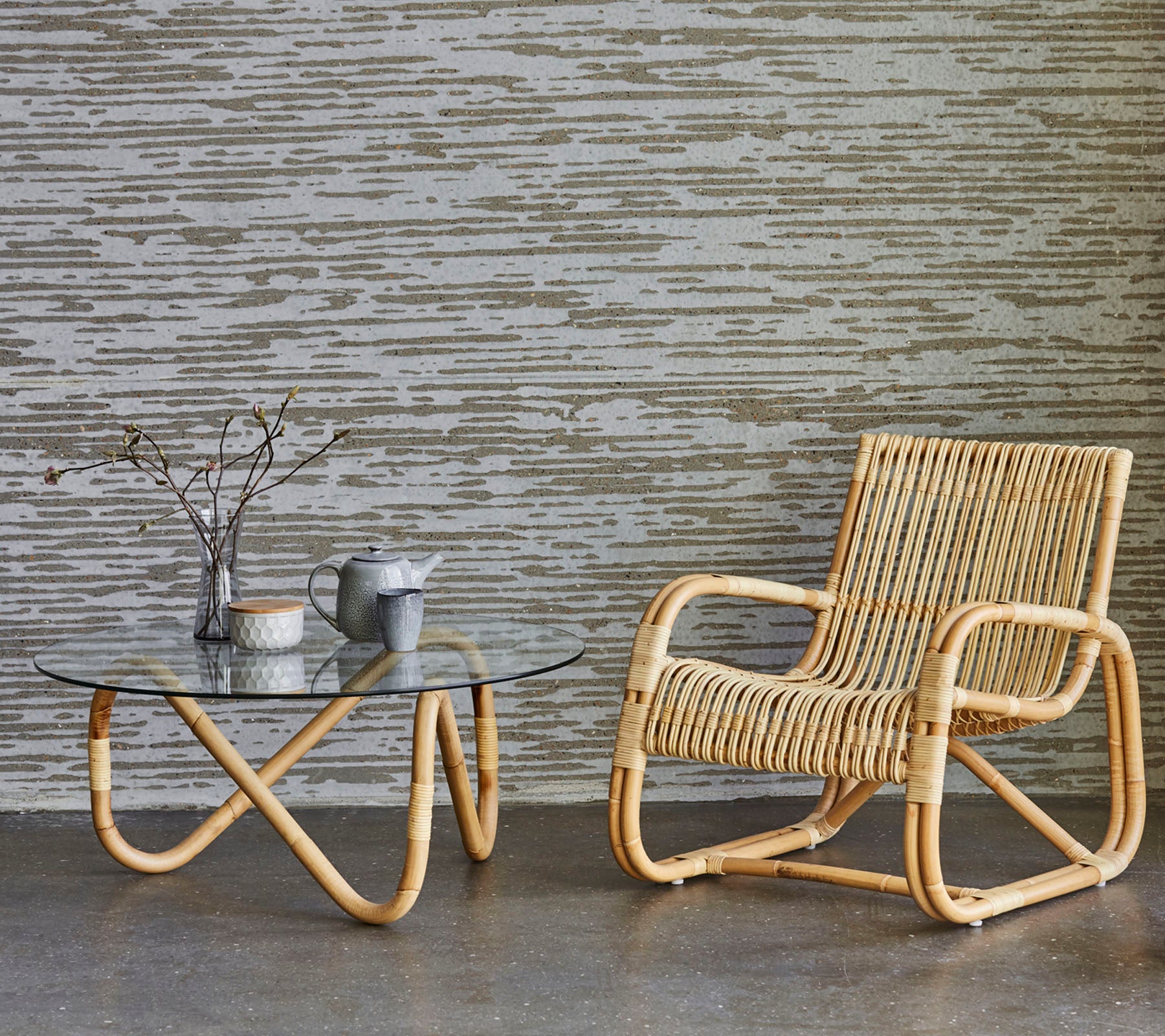 A stylish rattan chair next to a glass coffee table, featuring a vase with twigs and a small teapot, set against a textured background.