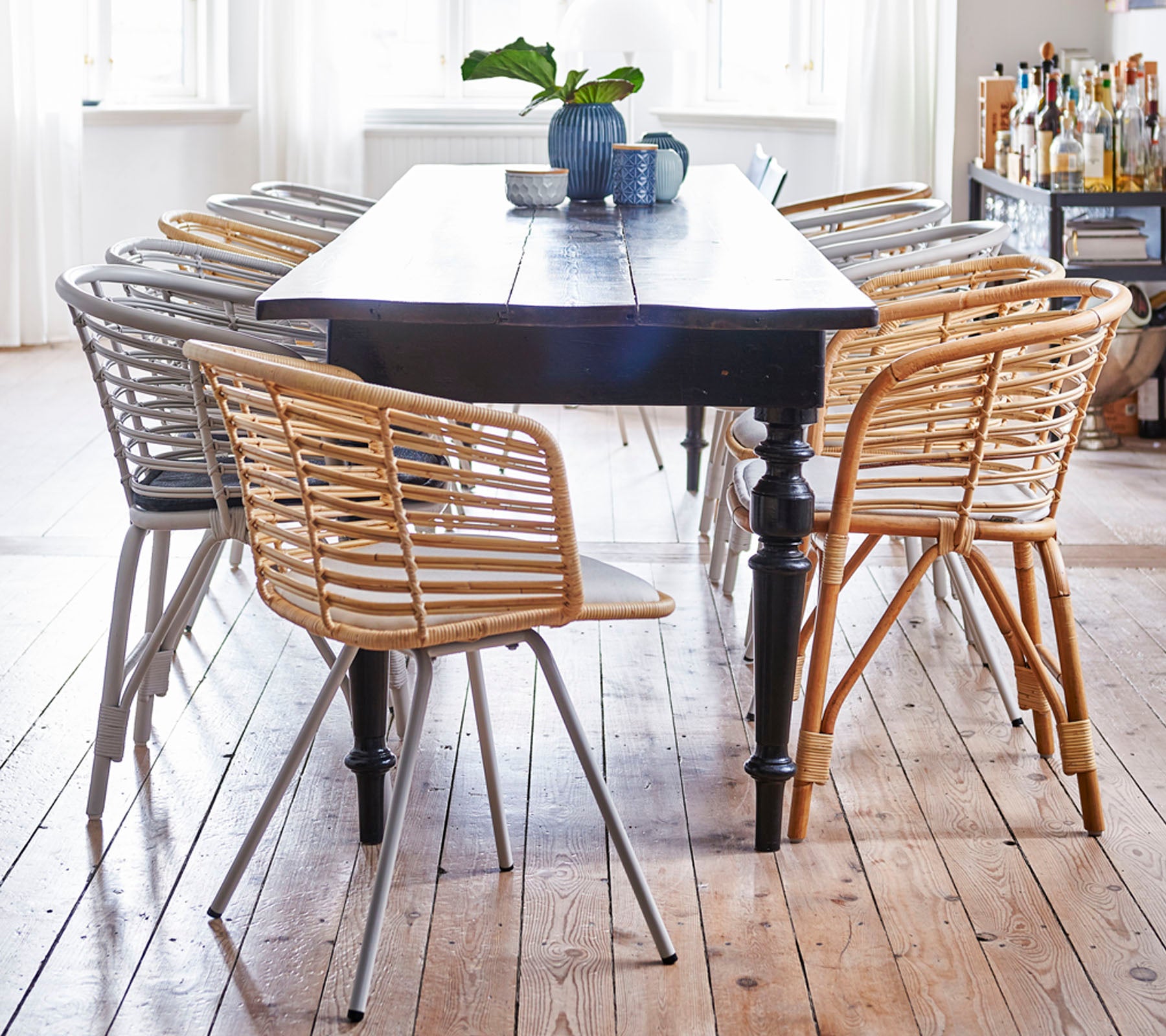 A wooden table surrounded by stylish, woven chairs in light and dark tones, with a small plant and a mug on top.