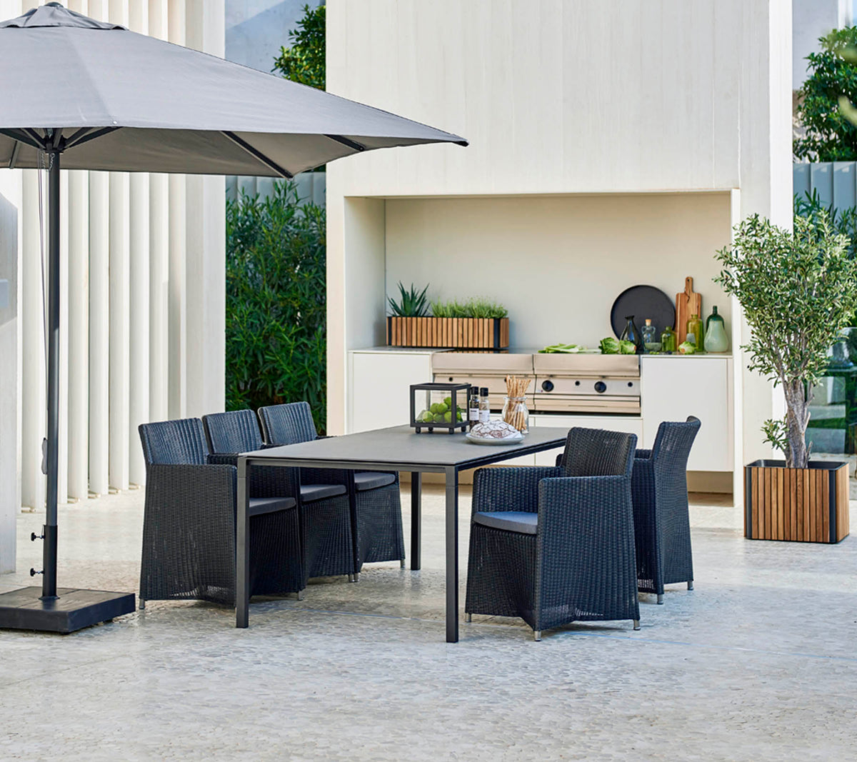 Modern outdoor dining set with a large table, six chairs, a stylish umbrella, and a backdrop featuring greenery and decorative elements.