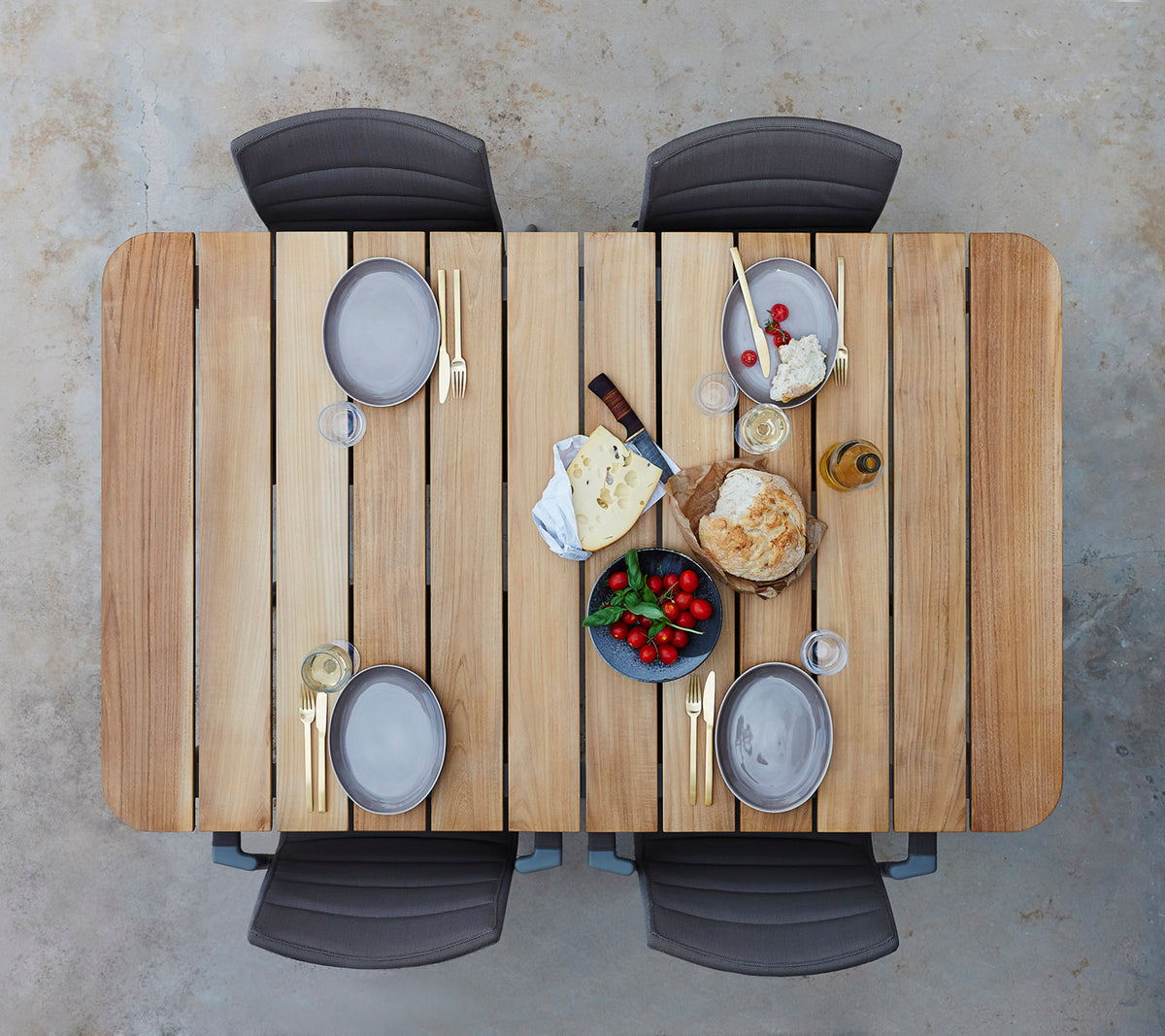 A wooden table set with plates, glasses, and an assortment of cheeses, fruits, and bread, surrounded by four chairs.