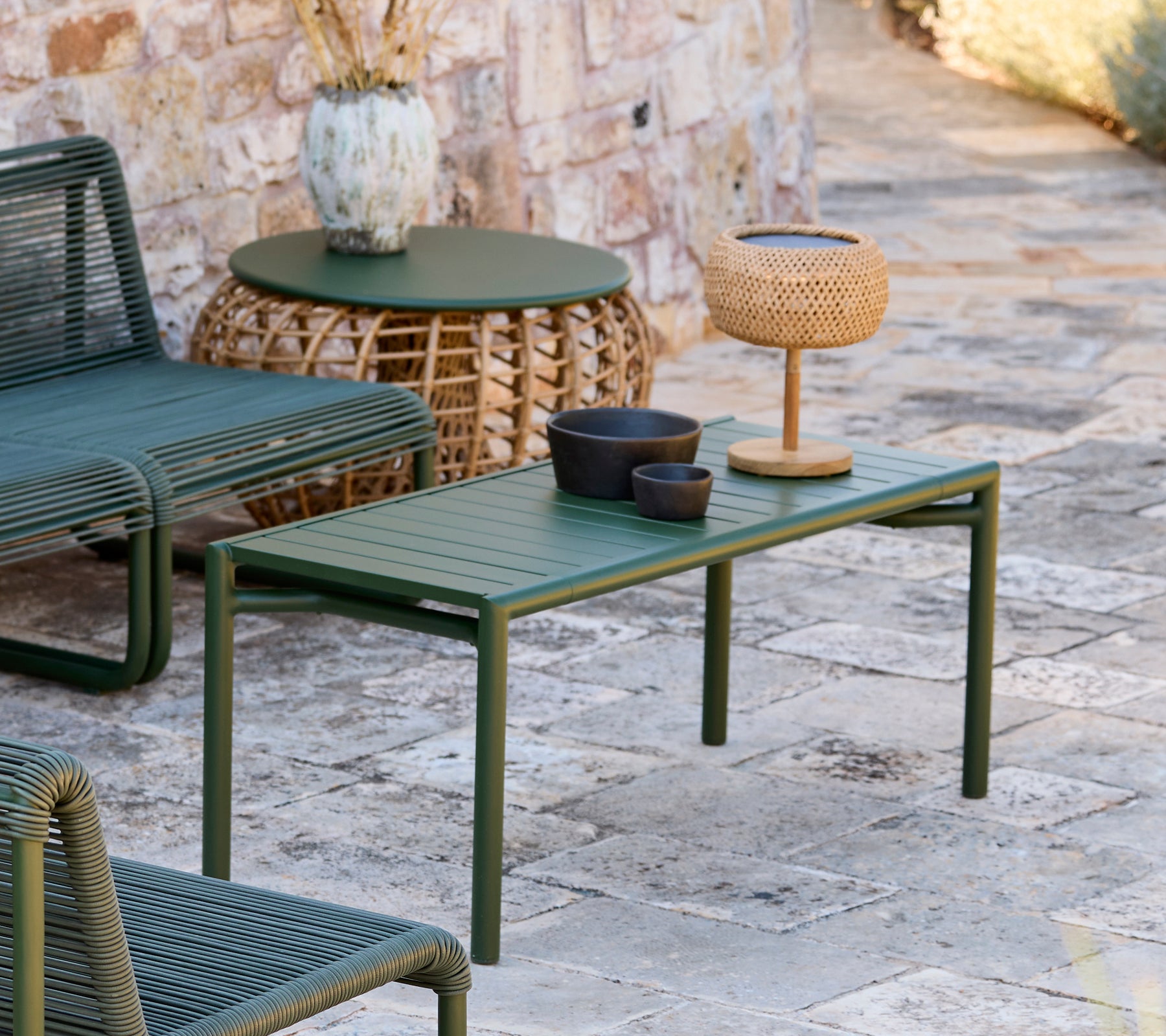 Green table with black bowl, brown vase, and woven lamp in a modern outdoor lounge area.