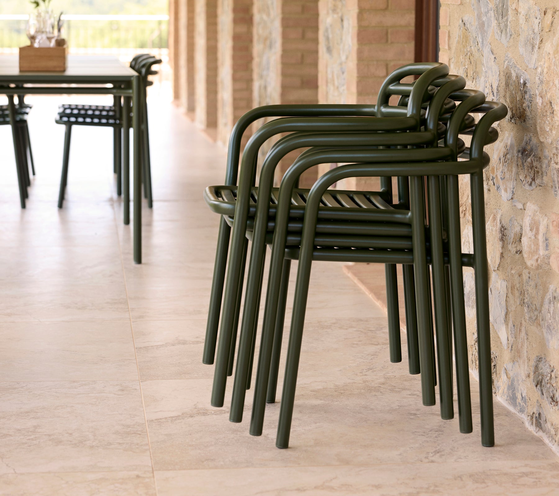 Stacked Bliss chairs in an outdoor setting, featuring a sleek design and earthy green color against a stone wall.