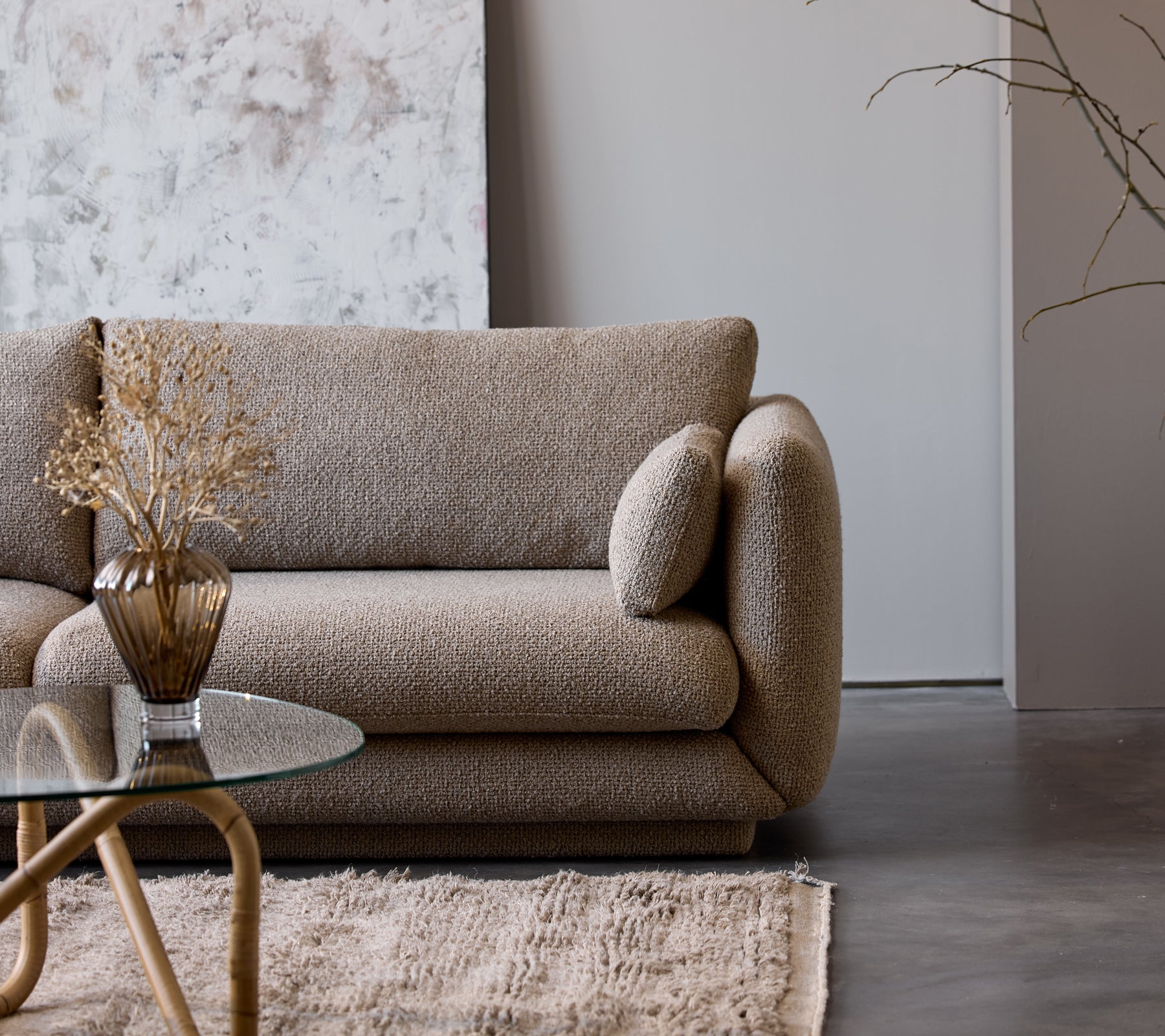 A taupe coffee table with a glass top sits next to a textured beige couch adorned with a decorative vase and dried flowers.