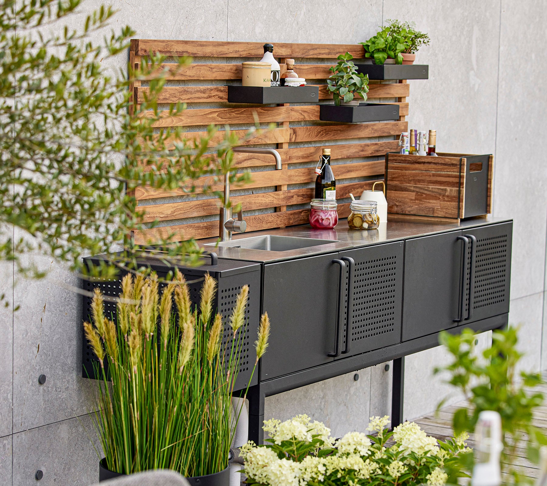 Modern black kitchen unit featuring a sink, wooden shelving, and various plants, creating a stylish and functional workspace.