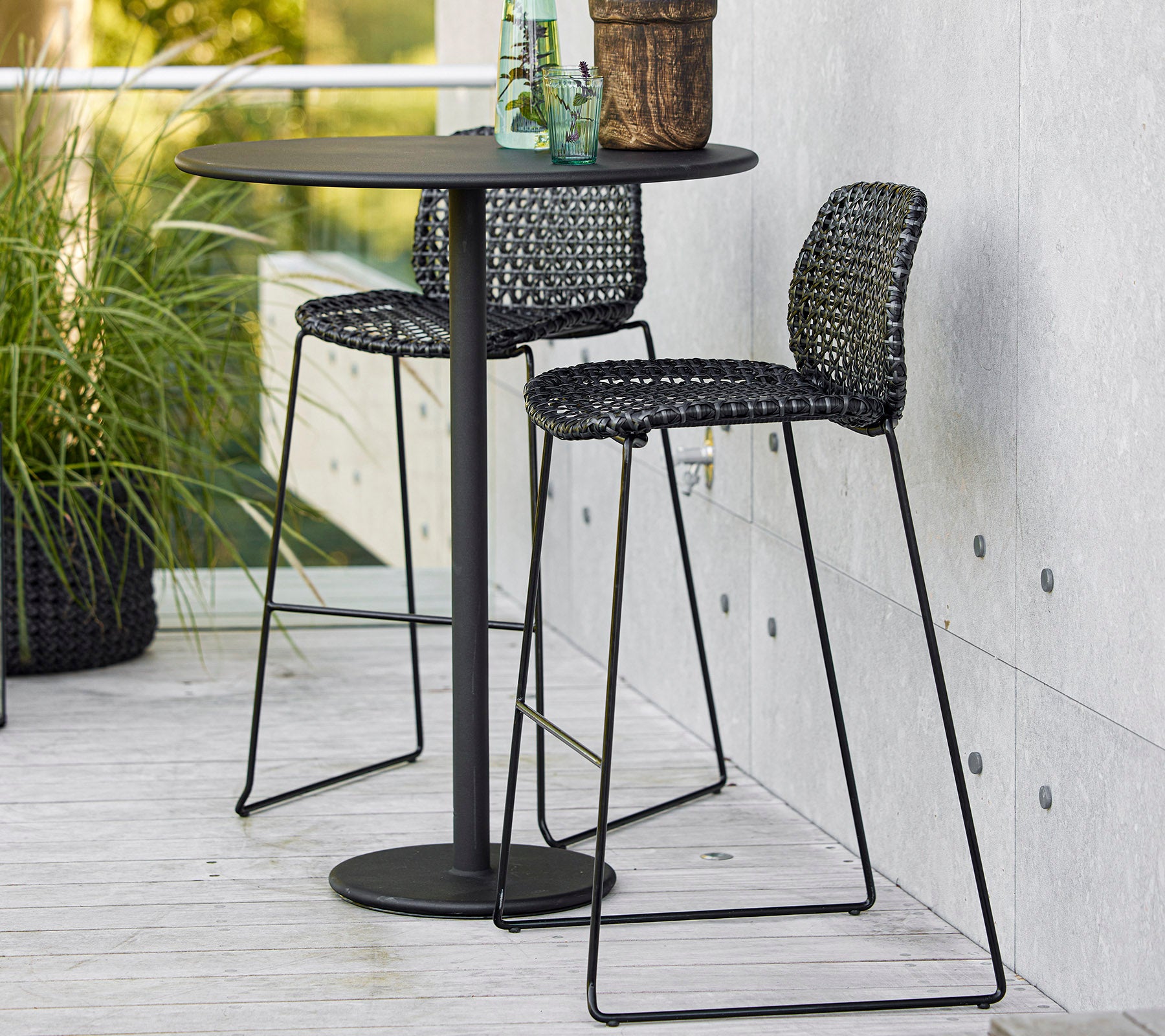 Stylish black bar table with two woven chairs and a potted plant, perfect for casual dining or relaxing.
