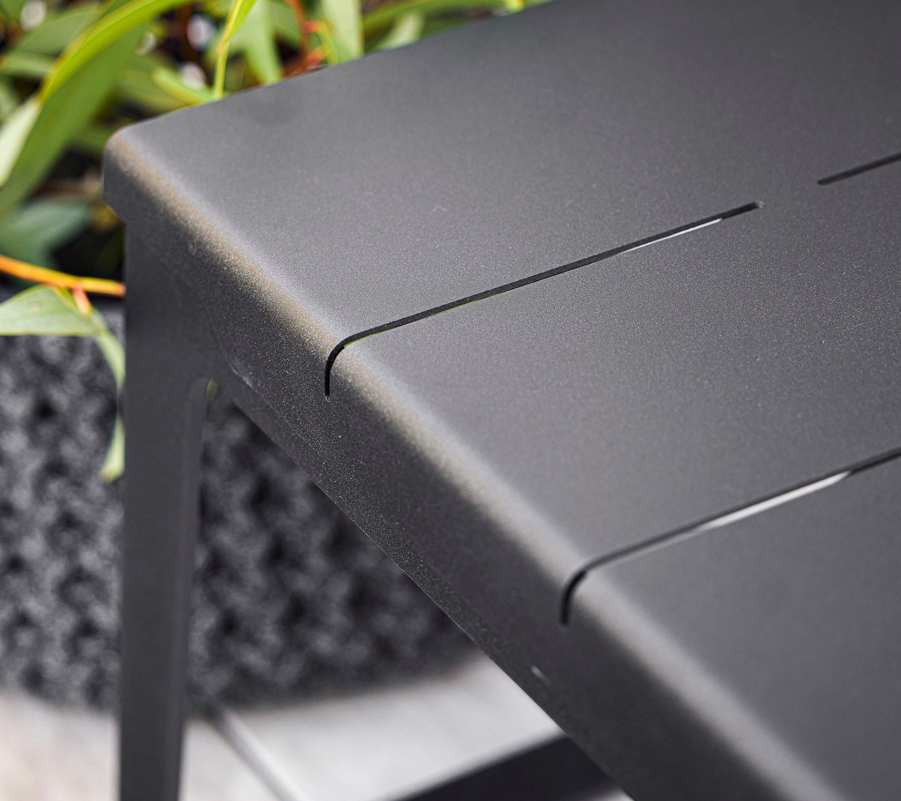 Black tabletop with subtle slits, featuring a minimalist design, complemented by greenery in the background. Ideal for modern spaces.