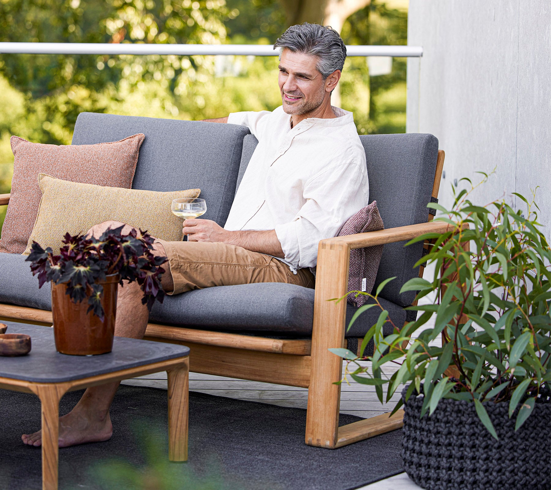 A man enjoying a drink while seated on a comfortable sofa, surrounded by plants and decorative pillows.