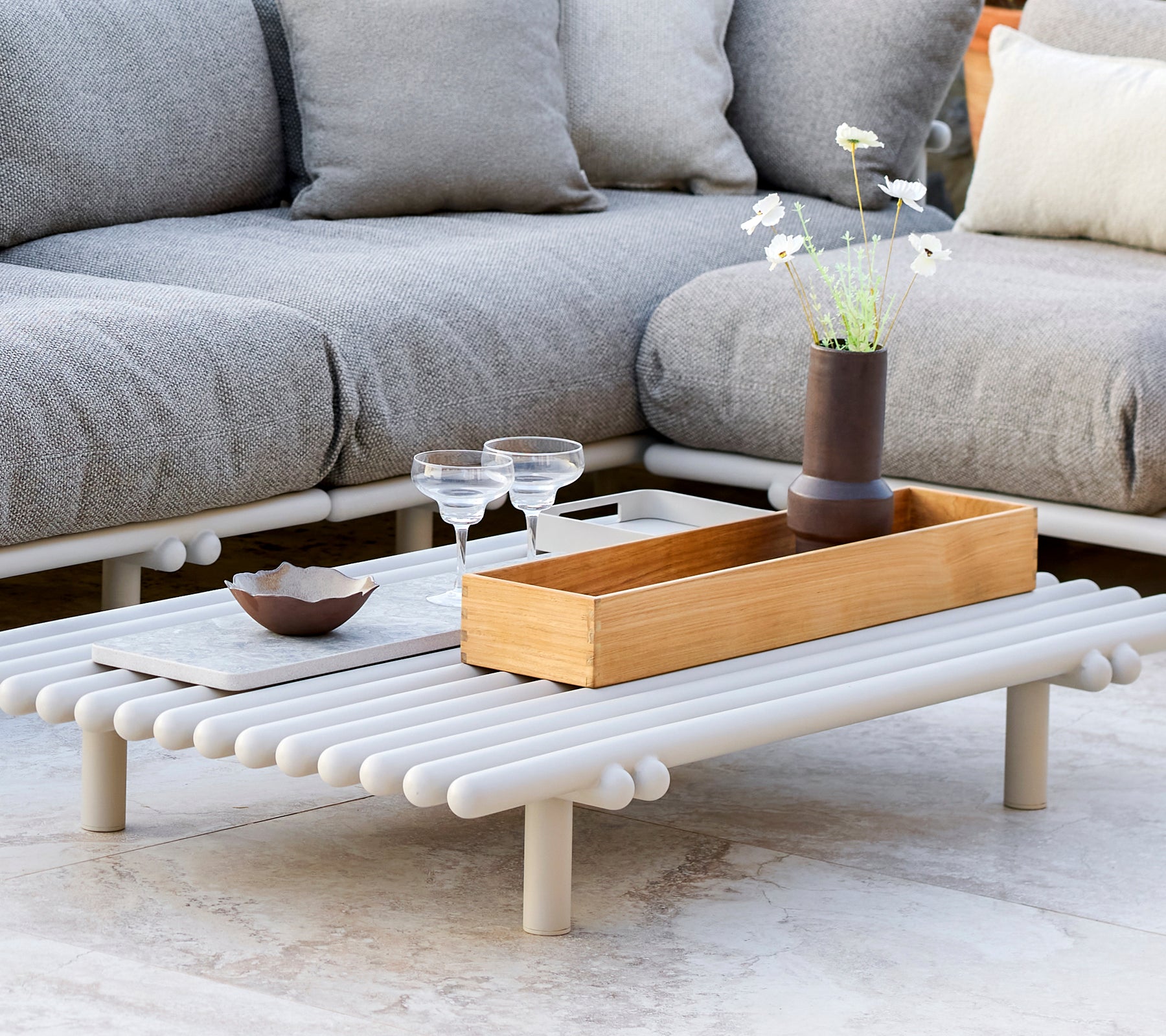 A stylish coffee table with a teak storage box, featuring a minimalist design and decorative elements like glasses and a small vase.