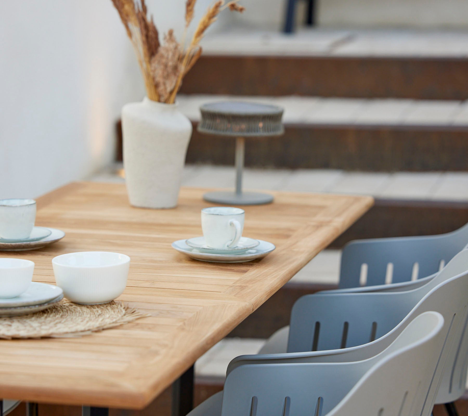 A wooden table set with cups and plates, decorated with a vase of dried flowers and grey chairs nearby.