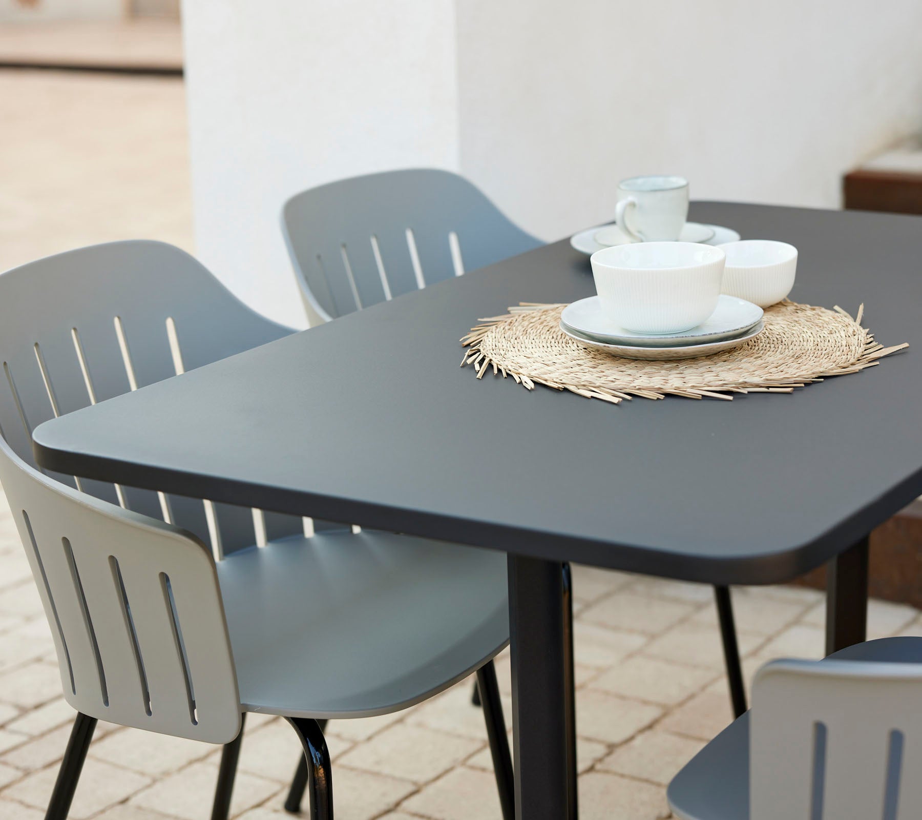 Modern cafe table set with a black tabletop, surrounded by grey chairs, featuring dishes and cups on a straw placemat.