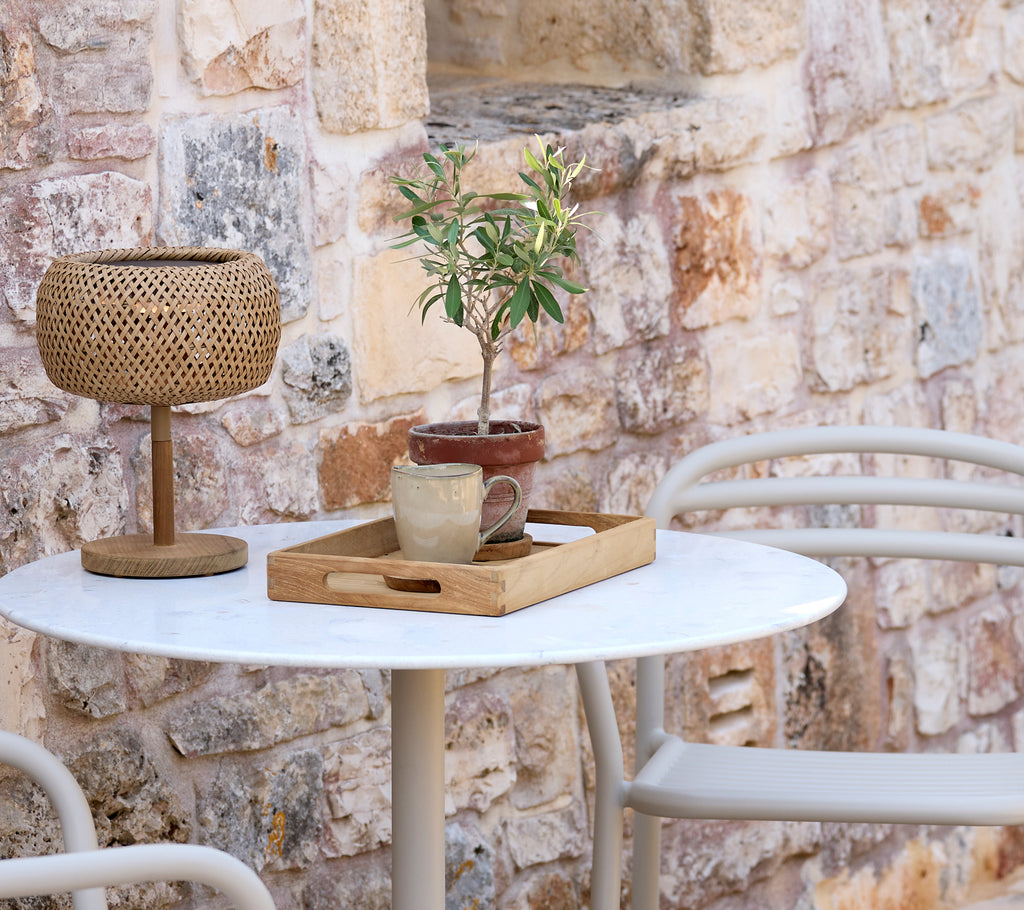 Modern outdoor setup featuring a white table, woven lamp, wooden tray, and a potted plant.