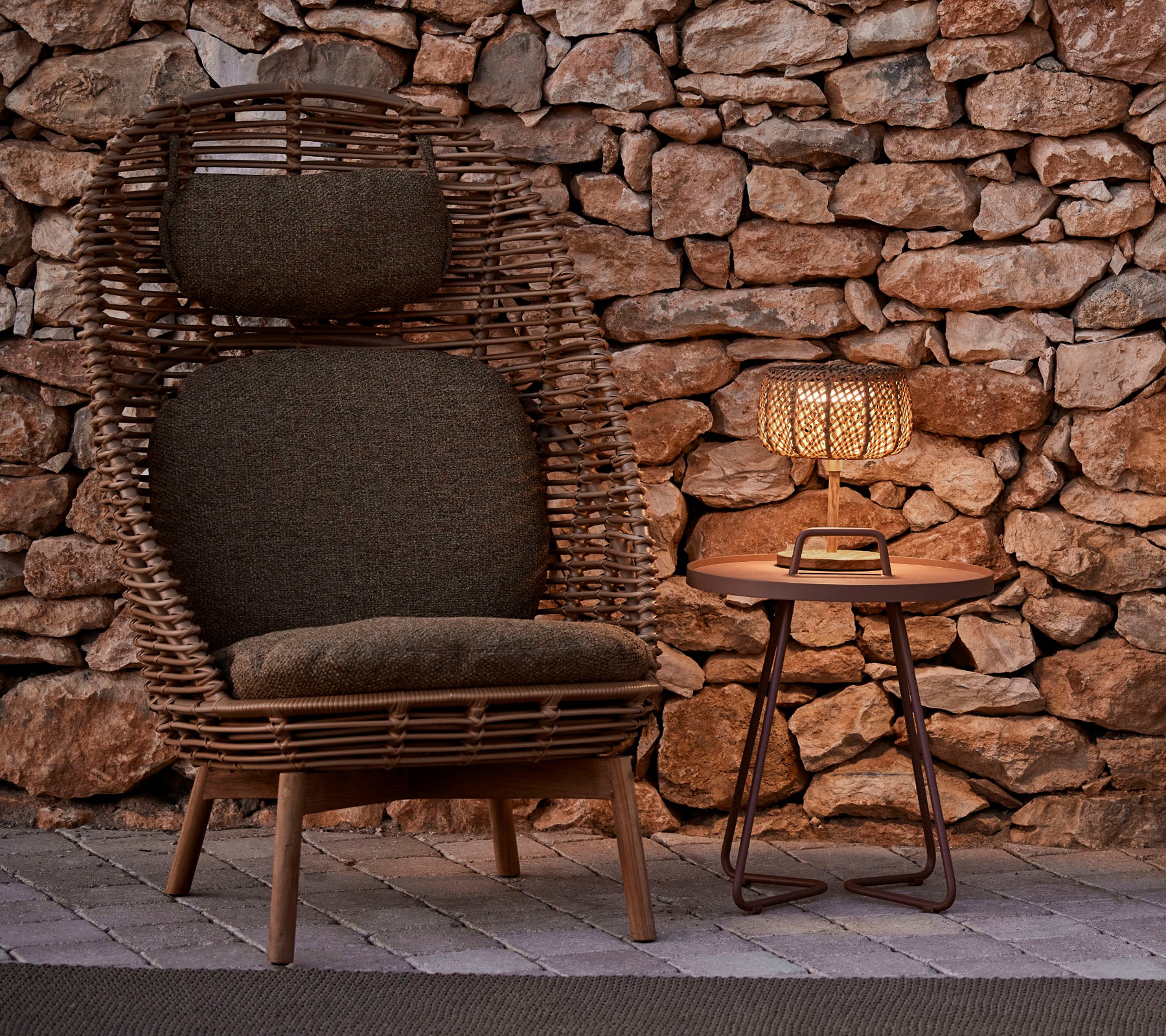 A stylish woven chair with cushions beside a wooden table and a glowing lamp, set against a rustic stone background.