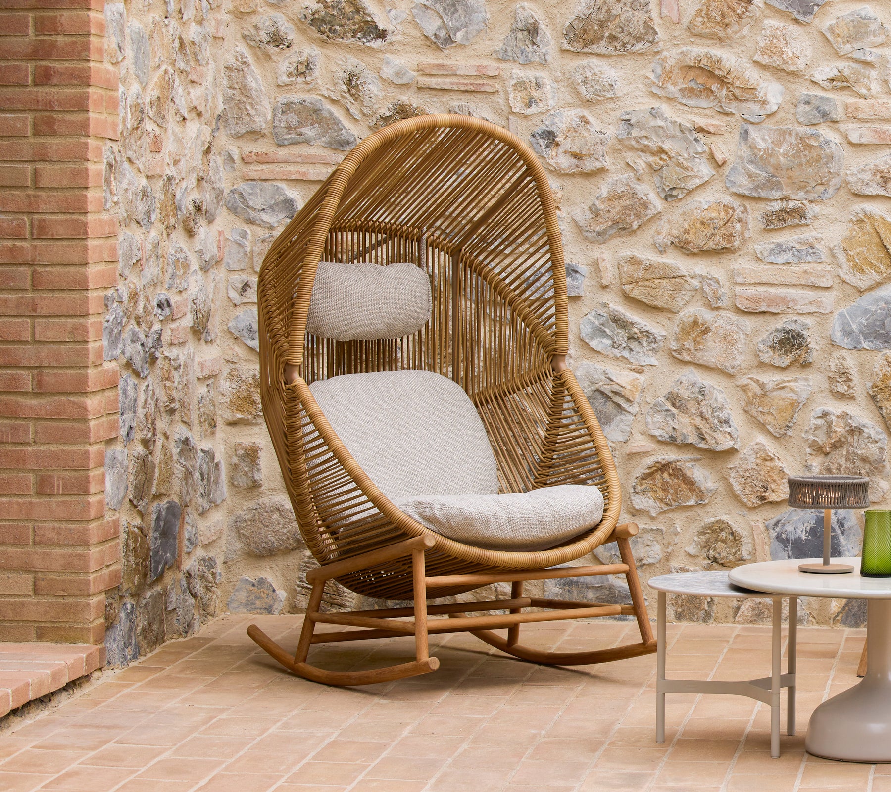 Hive rocking chair with soft cushions beside Glaze coffee tables, set against a textured stone wall.