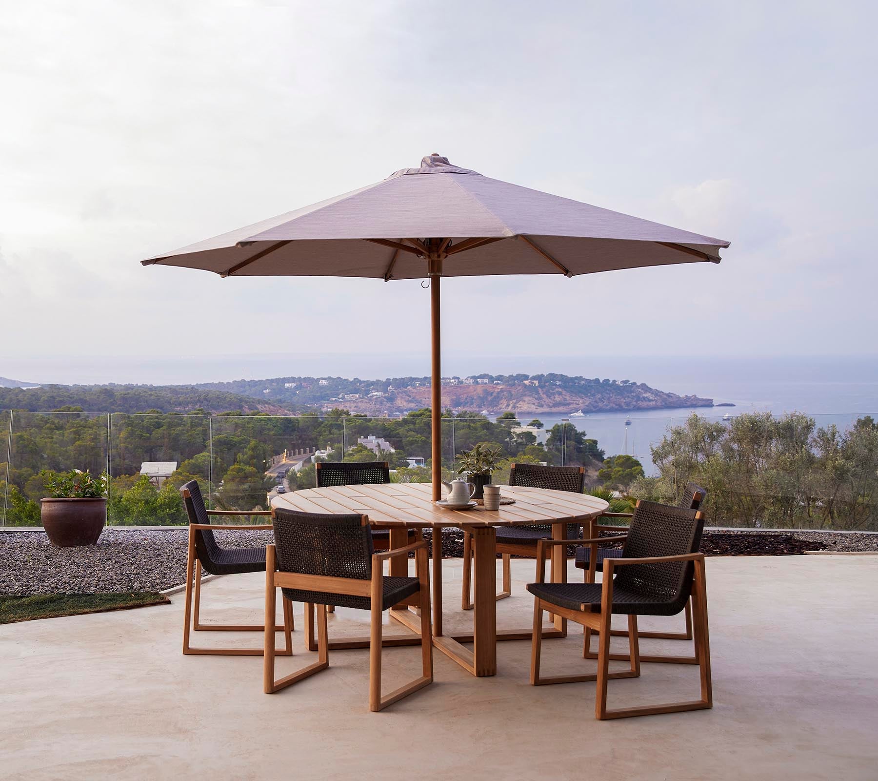 A round wooden table with six chairs under a large umbrella, set against a scenic backdrop of hills and the sea.
