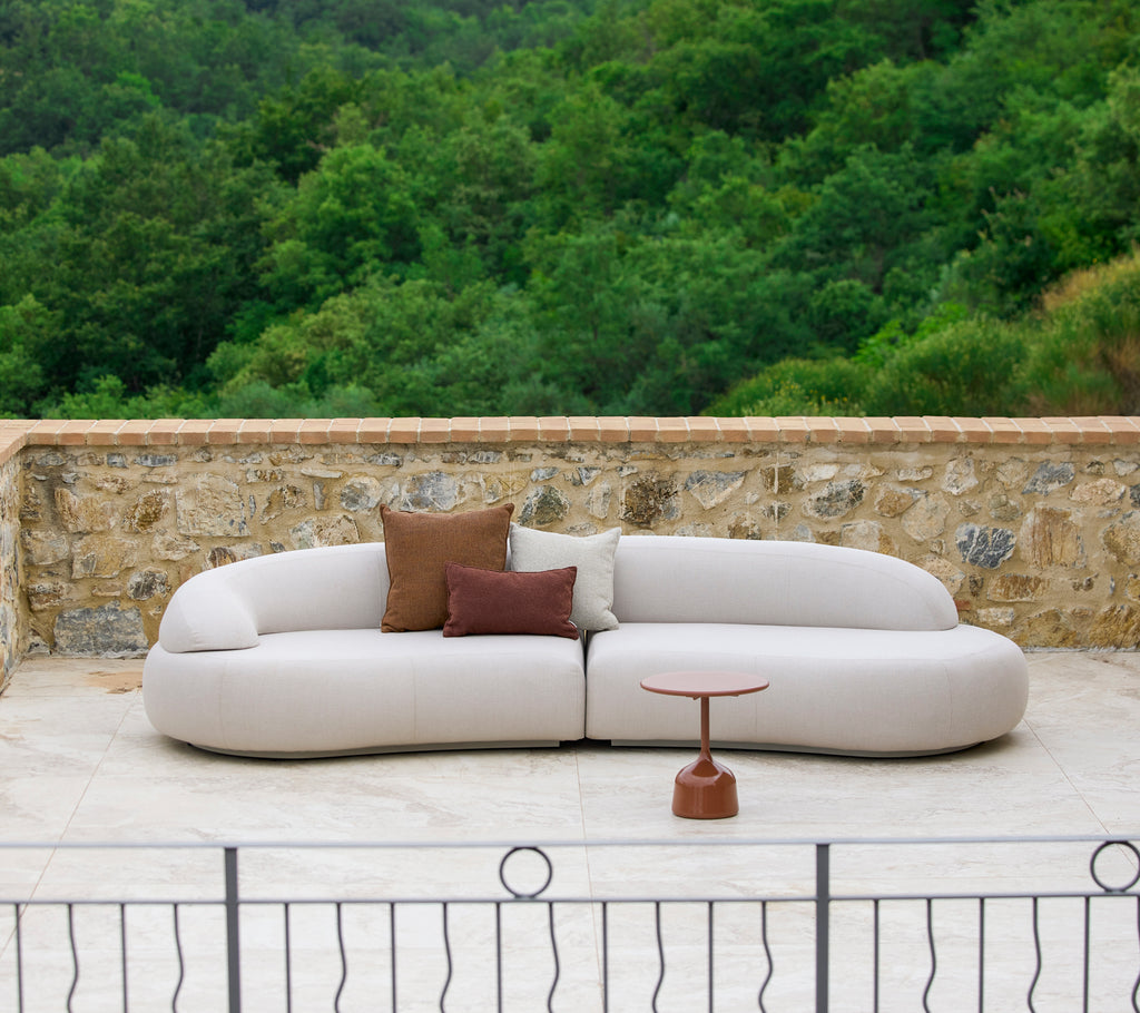 A modern, curved sofa with neutral upholstery and decorative pillows, complemented by a sleek side table in a natural setting.