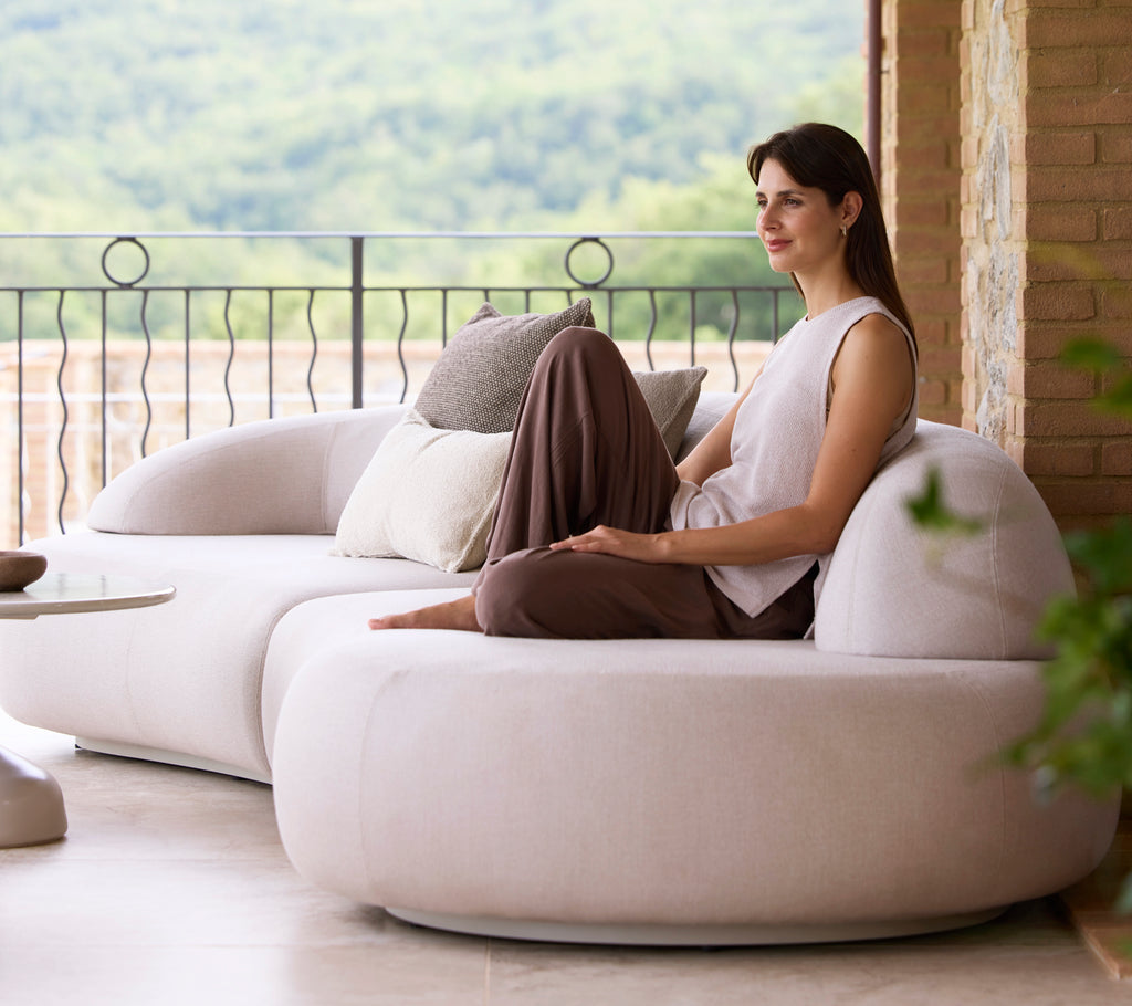 A woman sits comfortably on a sleek, modern sofa, accompanied by decorative cushions, enjoying a serene view of nature.
