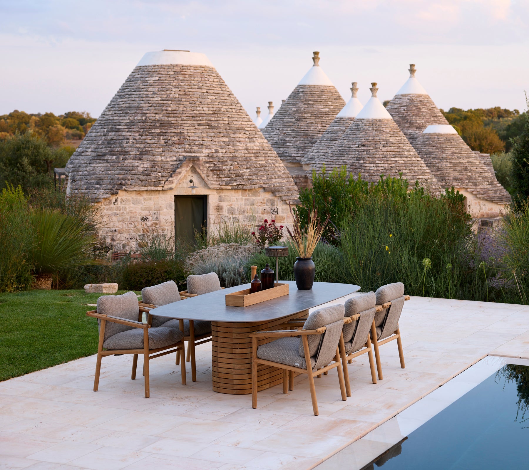 Grey dining chairs around a rectangular table near traditional trullo structures.