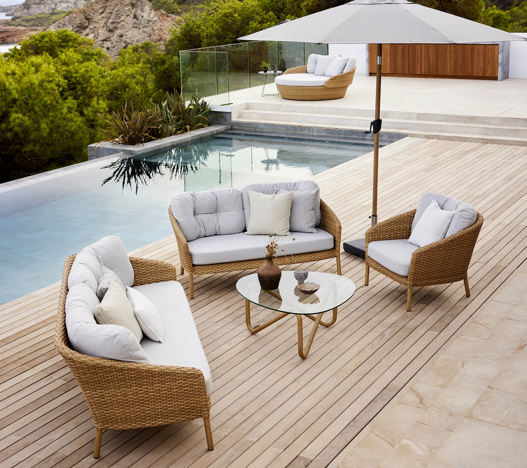 Modern outdoor lounge area featuring a glass table, comfortable seating, and an umbrella by a poolside with lush greenery.