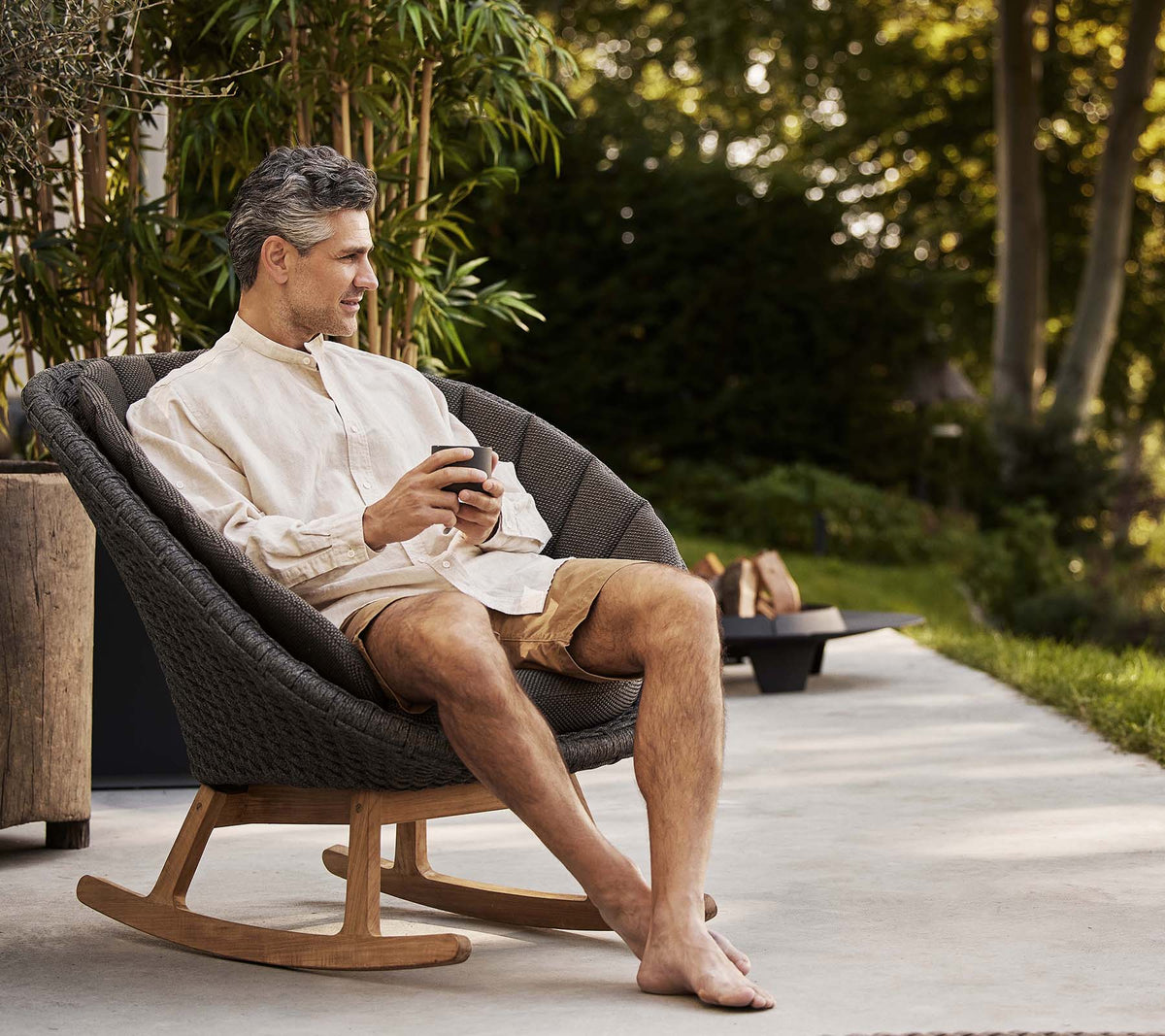 A man sits comfortably in a stylish peacock rocking chair, enjoying a moment of relaxation while holding a phone. Greenery is in the background.