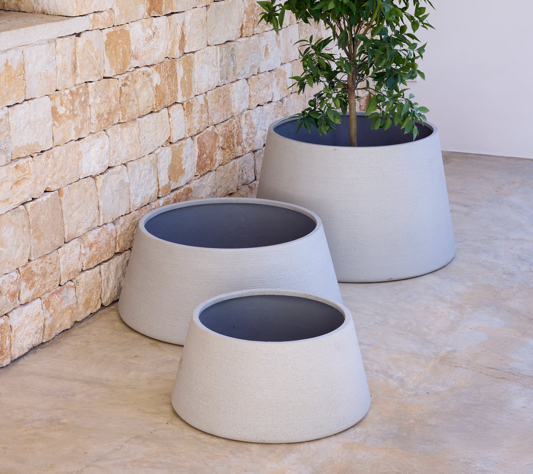 Three modern grey planters of varying sizes with a tree in one, against a stone wall.