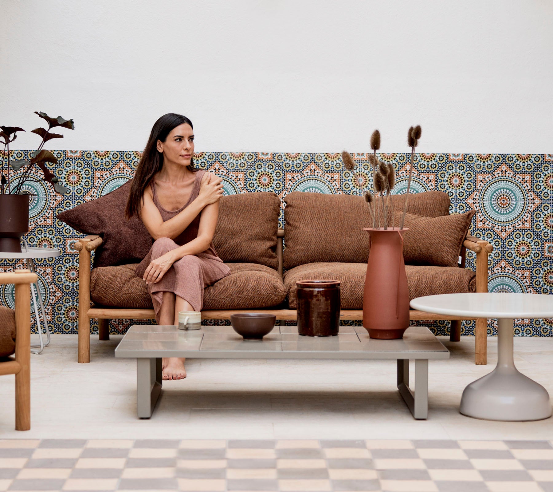A woman seated on a couch with decorative pillows, surrounded by vases and plants, against a patterned background.