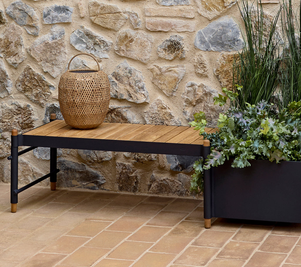 A stylish bench with a wooden top, accompanied by a woven basket and a decorative planter filled with greenery against a stone wall.