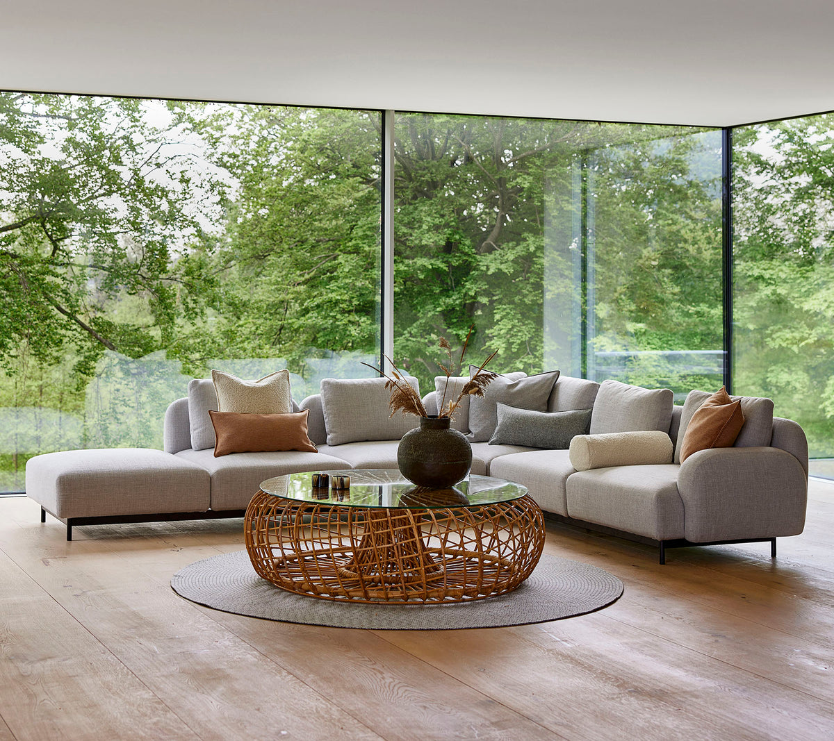 Modern lounge area featuring a modular sofa with decorative pillows, a round woven coffee table, and large glass windows showcasing greenery.