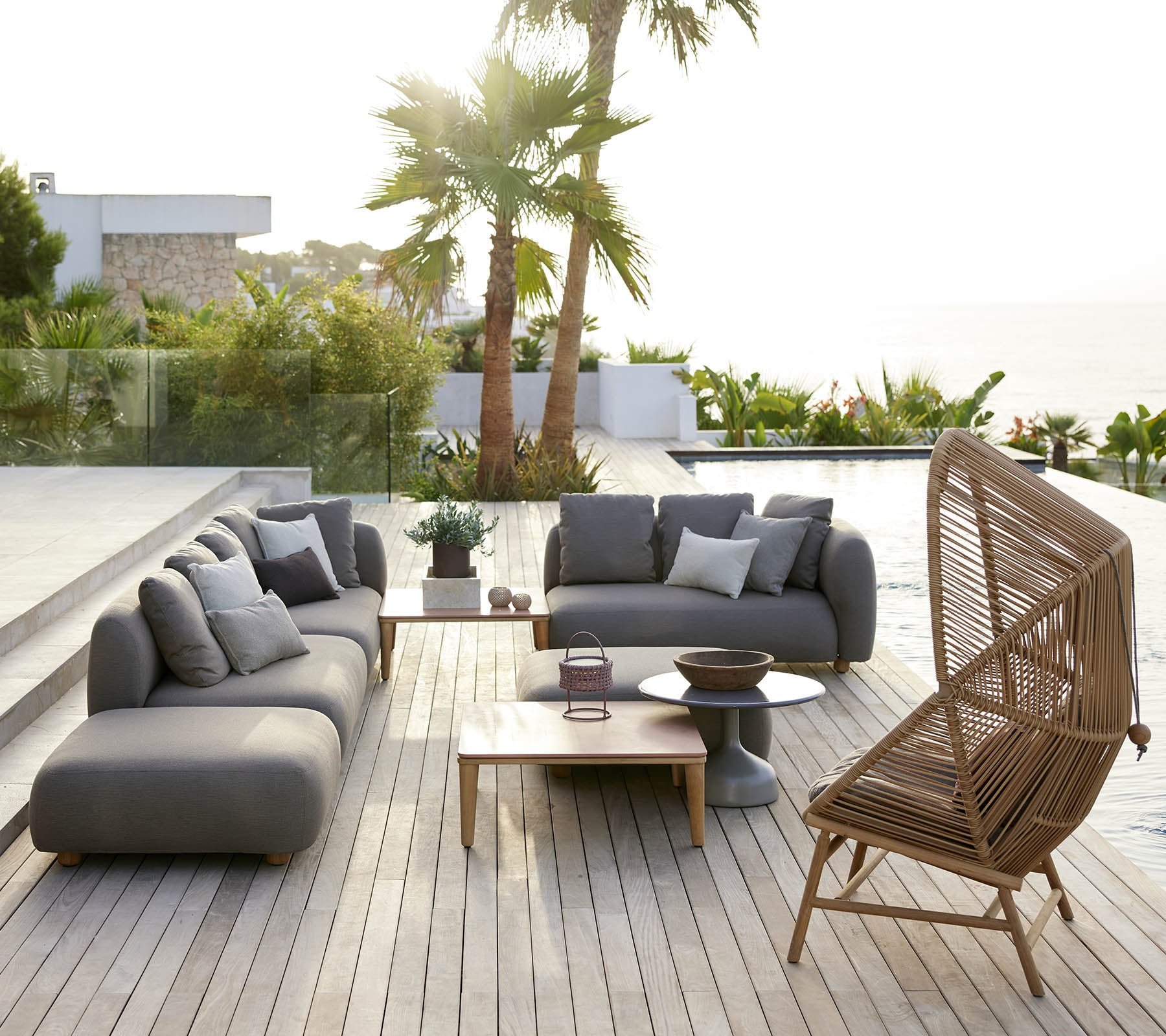 Stylish seating area featuring gray sofas and a wicker chair, complemented by a wooden table and lush greenery in the background.